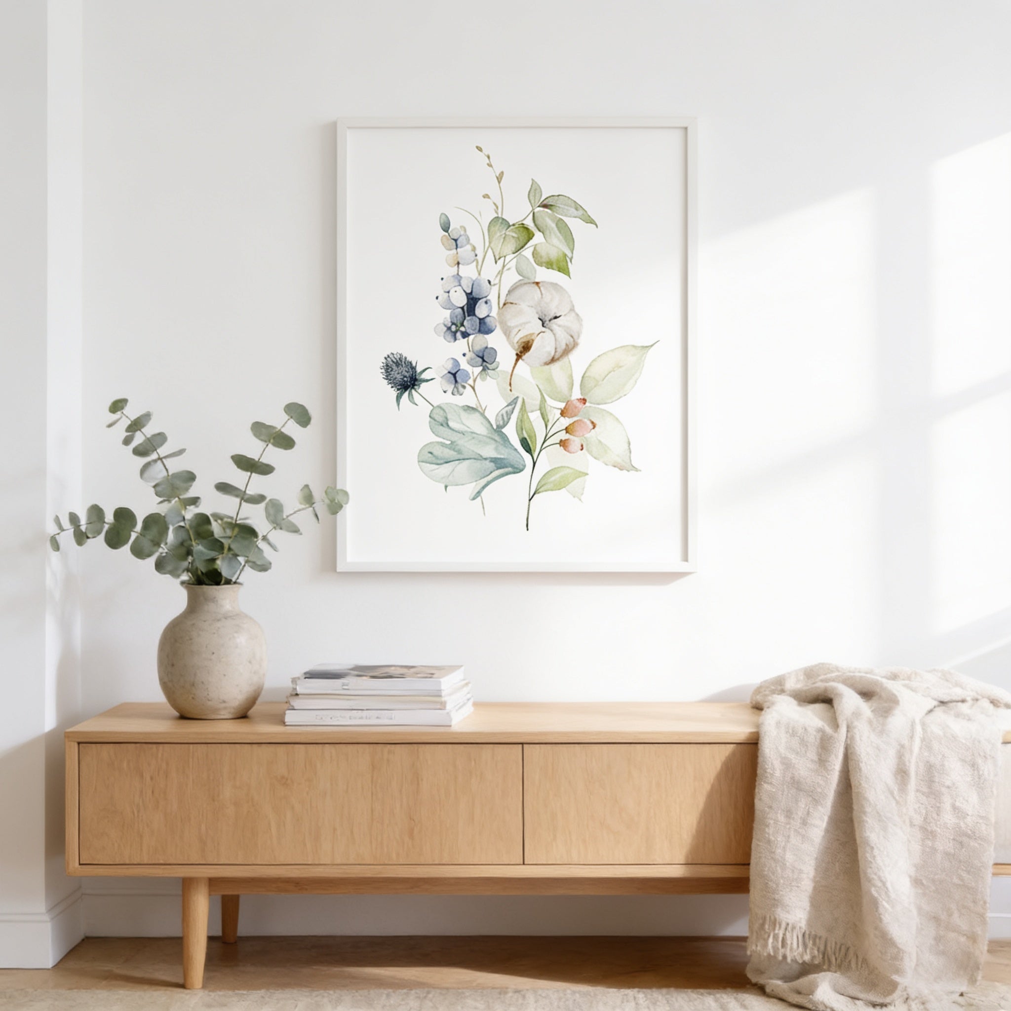a wooden console table with a vase of greenery and a stack of books on it, set against a white wall with a framed print of a floral arrangement hanging above.