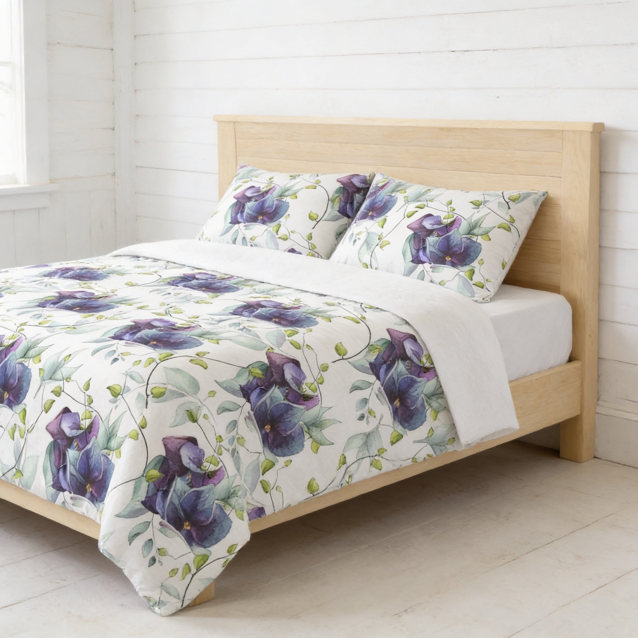 a bed with a white and purple floral comforter and matching pillows, set against a white wooden headboard and a white wall.