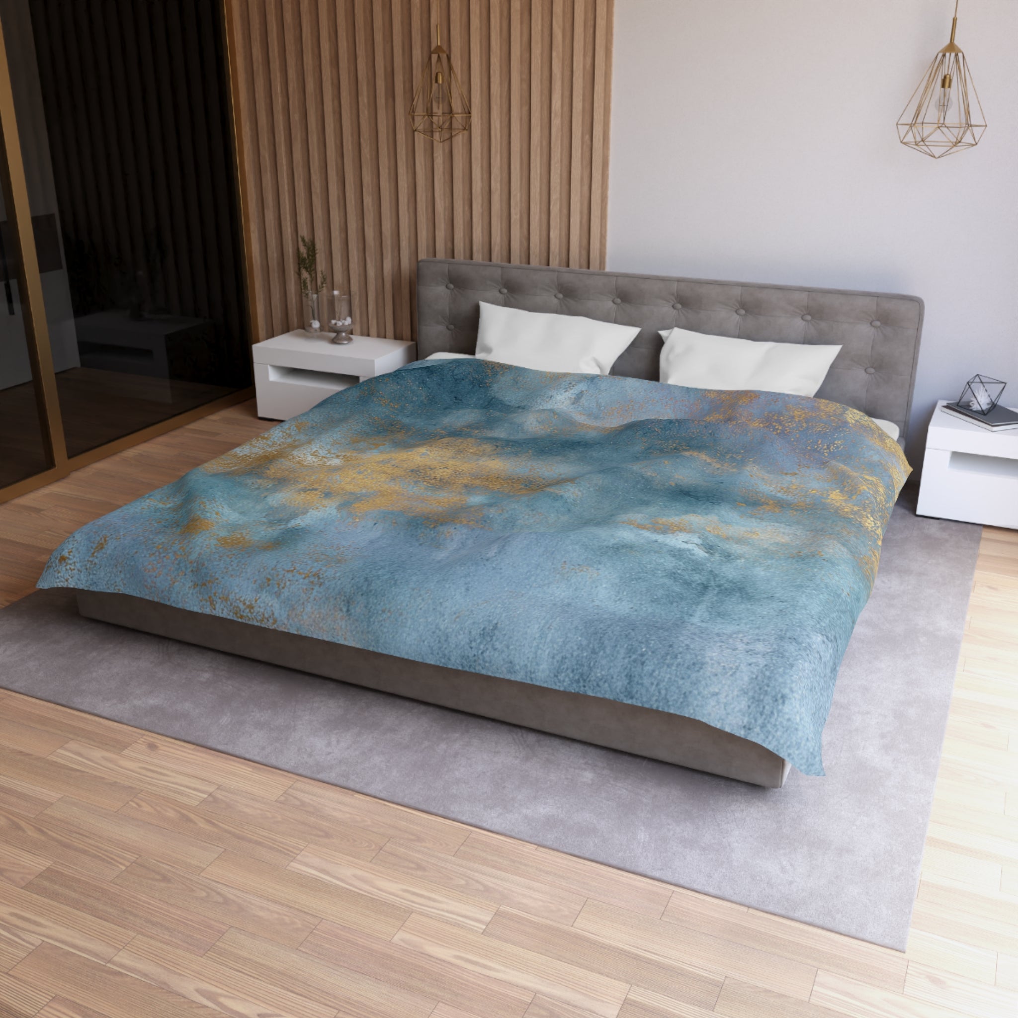 a modern bedroom with a large bed, a gray headboard, and a blue and gold patterned comforter. The room has a wooden floor and white nightstands on either side of the bed. There is a pendant light hanging above the bed, and a vase on one of the nightstands. The overall design and color scheme of the room create a serene and stylish atmosphere.