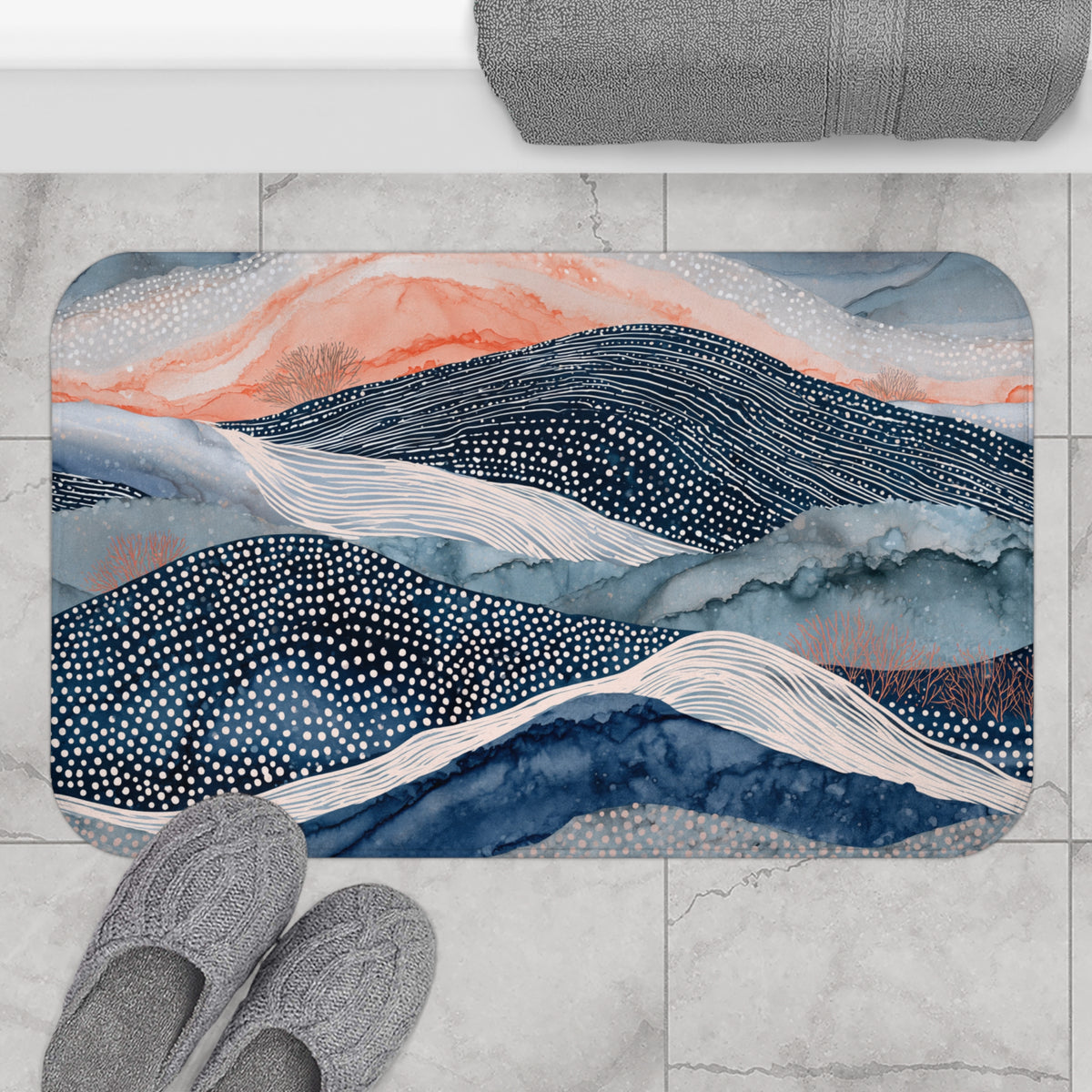 a bathroom rug with a mountain landscape design, including hills, valleys, and a body of water. The rug is placed on a tiled floor, and there are gray slippers visible in the foreground.