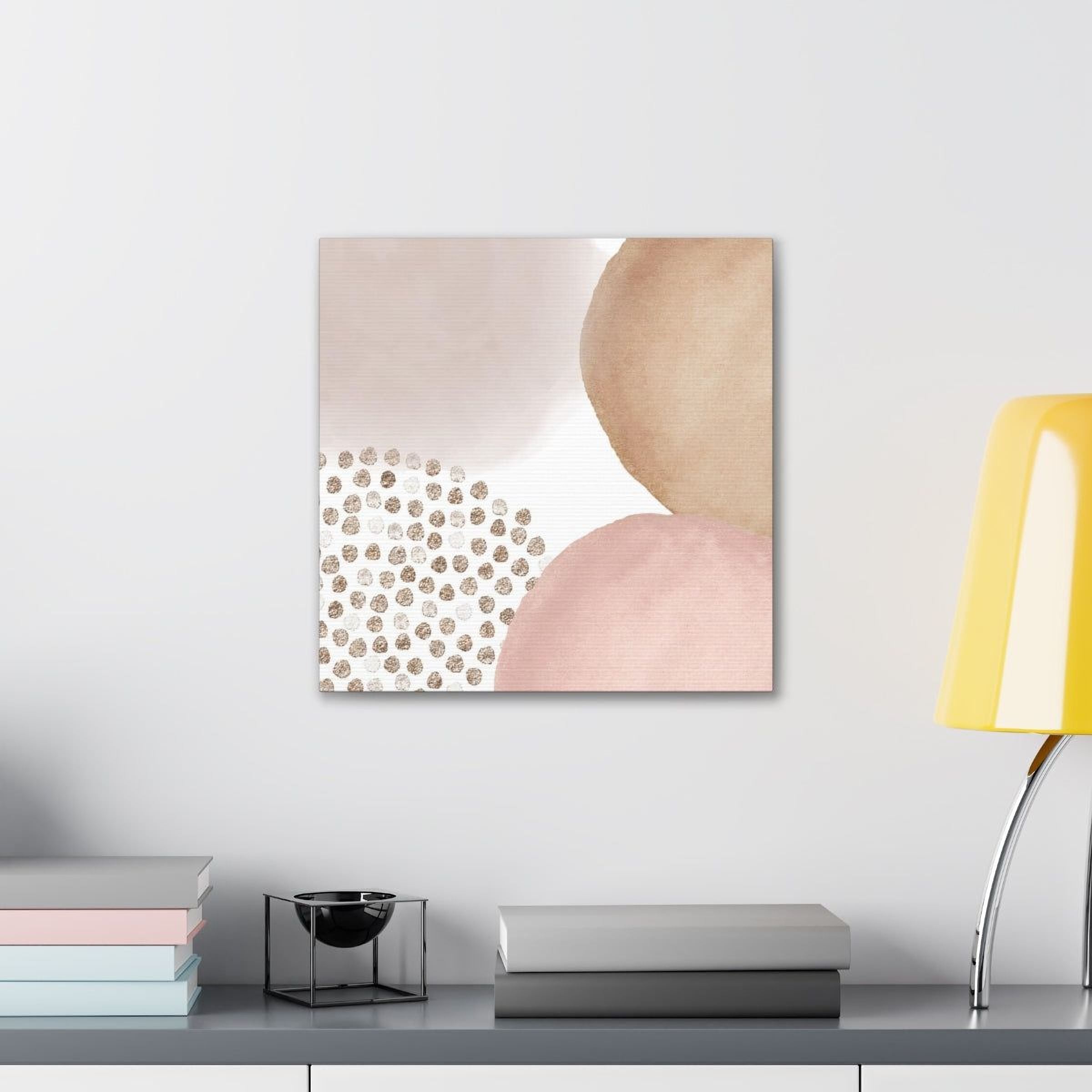 a minimalist and modern living space with a white wall, a shelf holding books, and a lamp. The focal point is a square canvas print with abstract shapes and dots in shades of pink and beige.