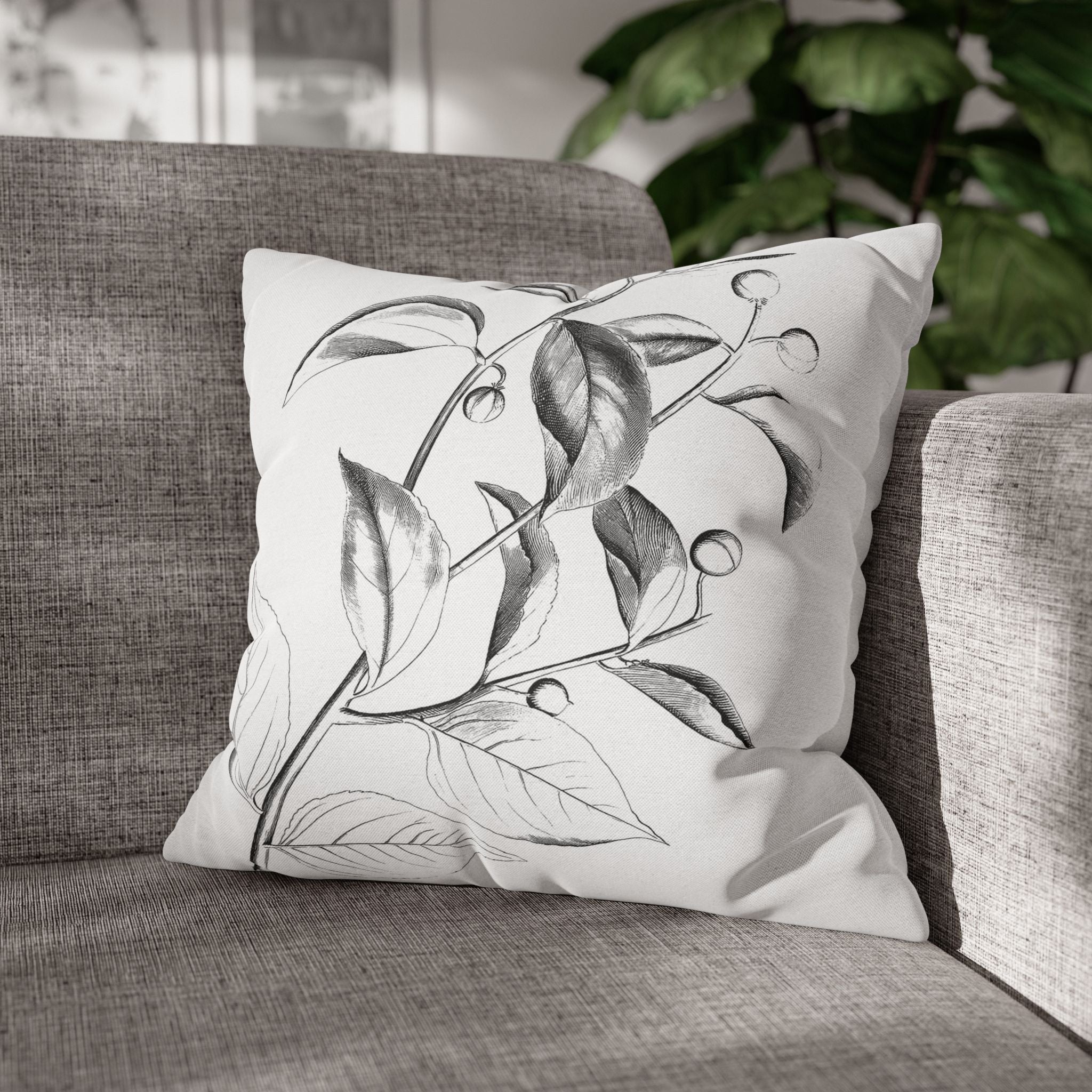 A white throw pillow with a black and white illustration of leaves and branches sits on a gray couch.