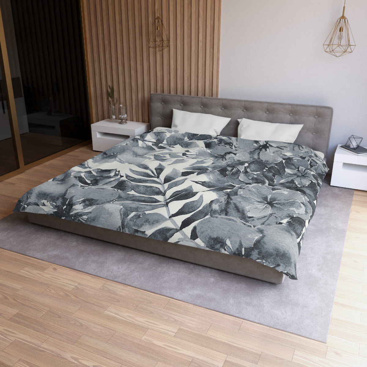 a modern bedroom with a large bed covered in a gray and white floral comforter. The bed is positioned against a wall with a wooden panel. The room also features a nightstand, a pendant light, and a rug on the floor.