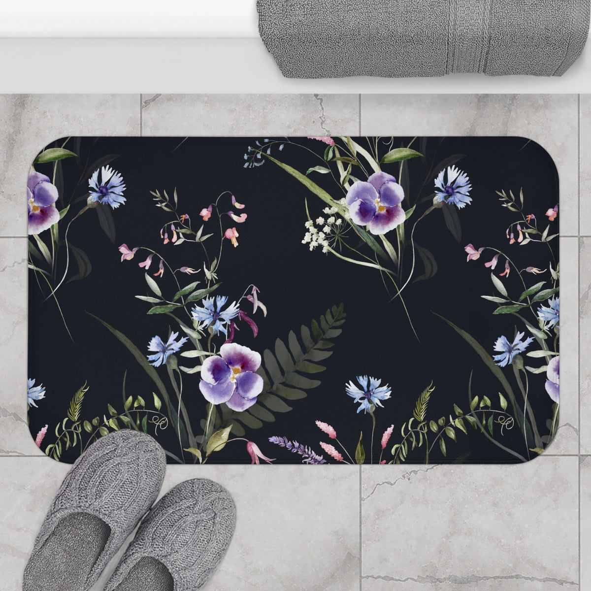 A bathroom mat with a floral pattern of purple and blue flowers, along with green leaves, is placed on a tiled floor. There are also gray slippers visible in the bottom left corner of the image.