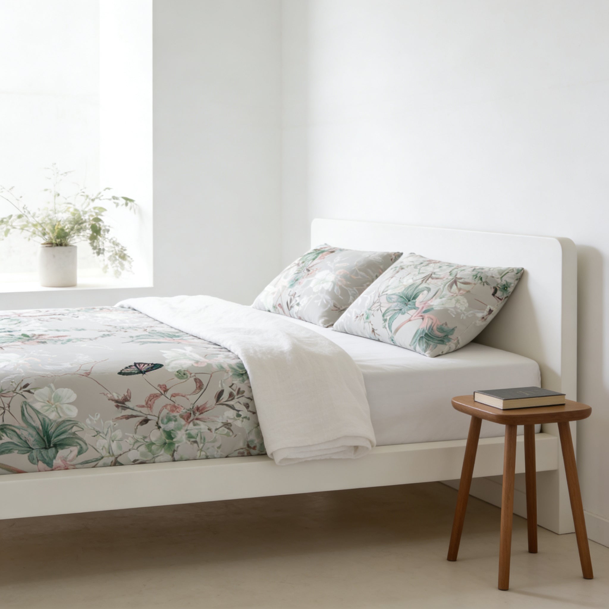 a serene and cozy bedroom scene, featuring a white bed with a floral bedspread and matching pillows, a small wooden side table, and a window with a plant on the sill.