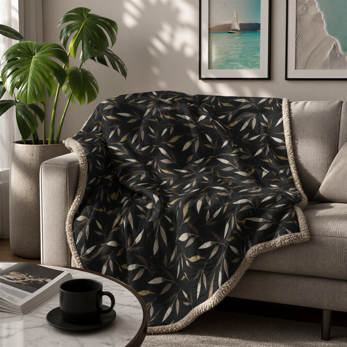 a cozy living room with a black and gold leaf-patterned throw blanket draped over a beige sofa. A coffee table with a black mug and a book is situated in front of the sofa. The room is decorated with two framed pictures on the wall, and a potted plant is placed on the floor.