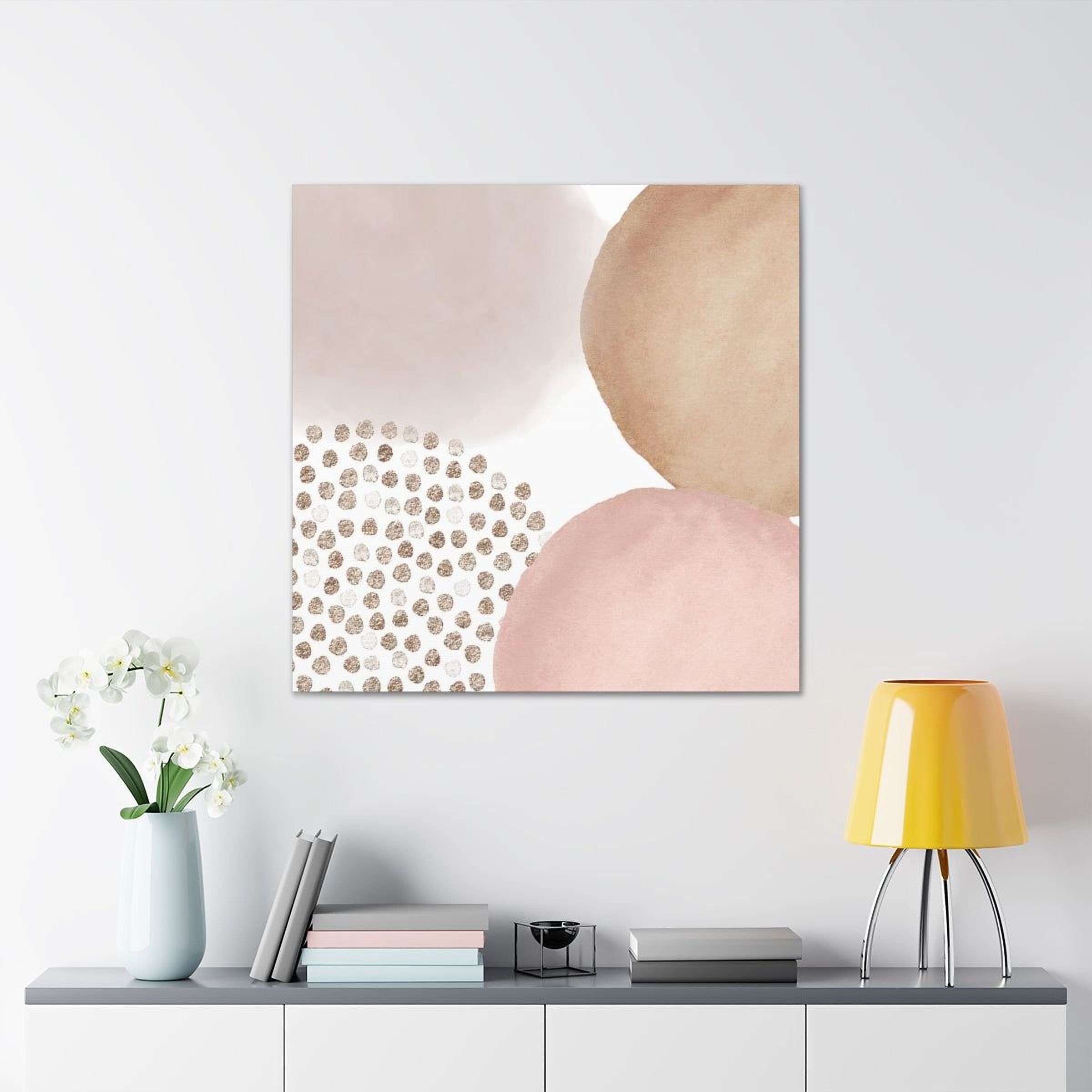 a minimalist living space with a white wall, a shelf with a vase of white flowers, a stack of books, and a yellow lamp. Above the shelf, there is a square canvas print with abstract shapes in shades of pink and brown, along with a pattern of small dots.