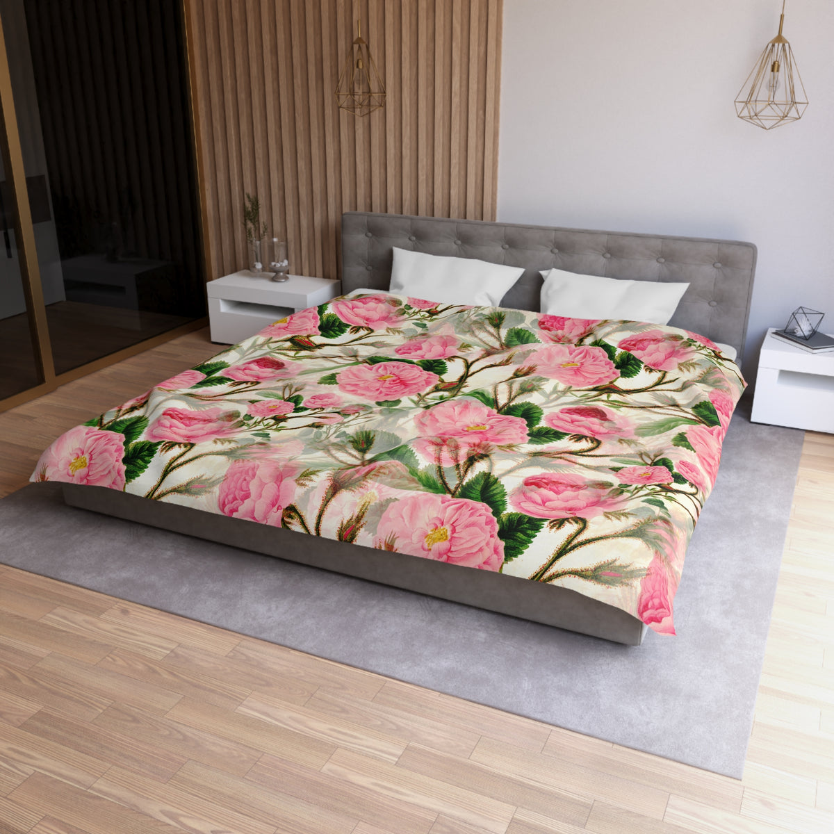 a bedroom with a large bed covered in a floral comforter featuring pink flowers and green leaves. The bed is positioned against a wall with a wooden panel. The room has a gray headboard and a white nightstand on each side of the bed. There is a pendant light hanging above the bed, and a vase is visible on a surface nearby.