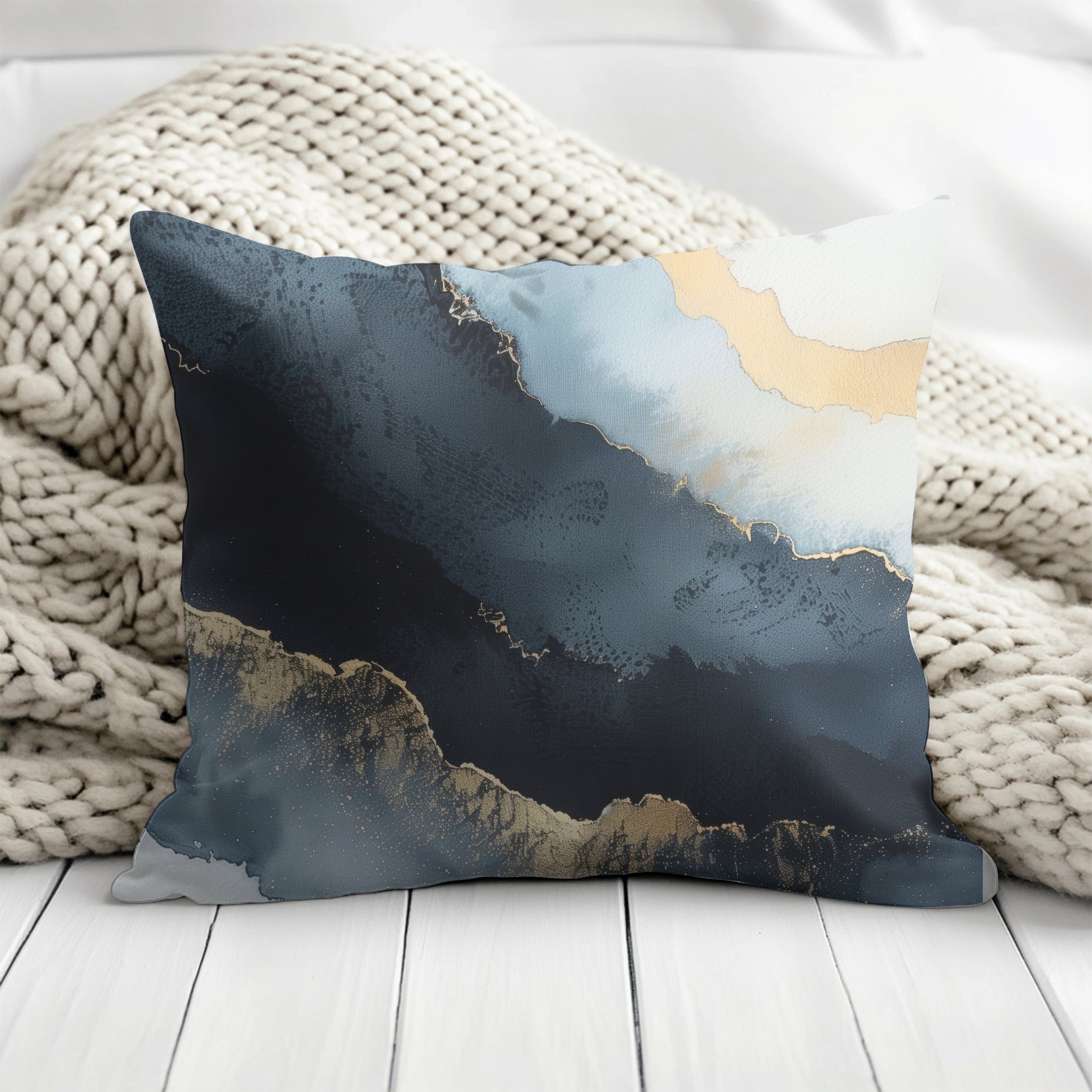 A decorative throw pillow with a striking abstract design in shades of blue and gold sits on a white wooden surface, partially covered by a cozy white blanket.