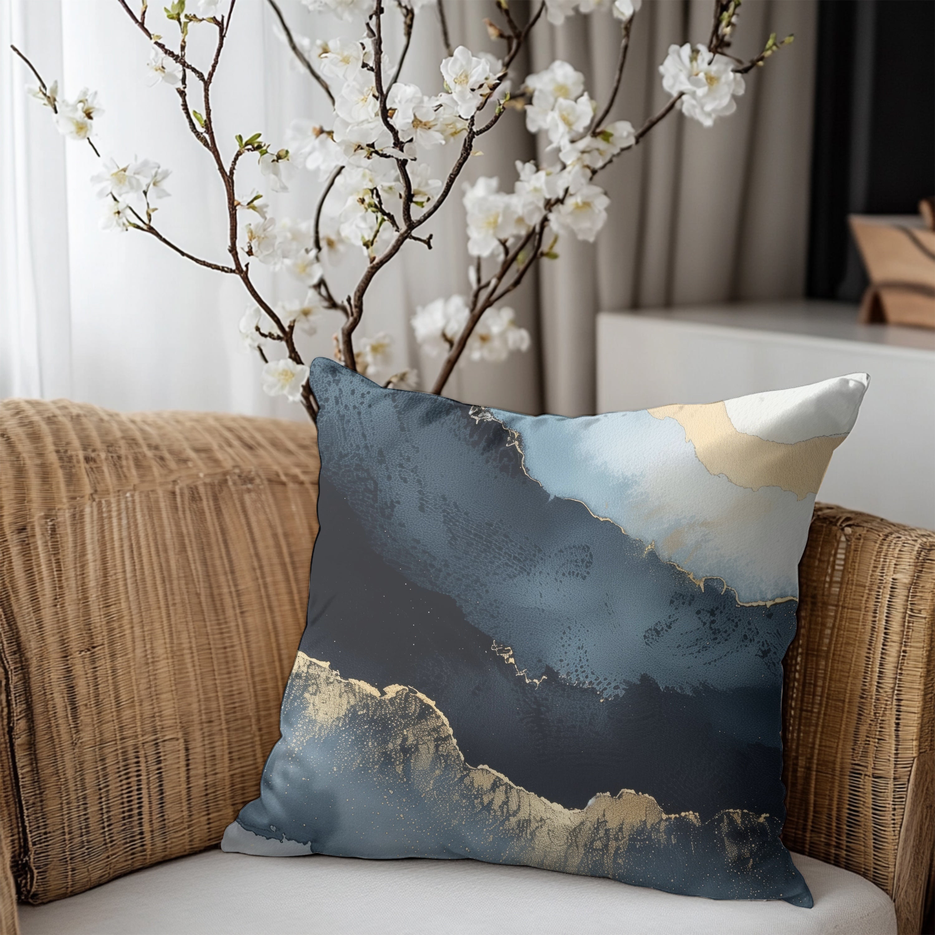 a decorative pillow with a blue and white abstract design, placed on a wicker chair. The pillow is positioned in front of a window with white curtains, and there is a branch of white flowers visible outside.
