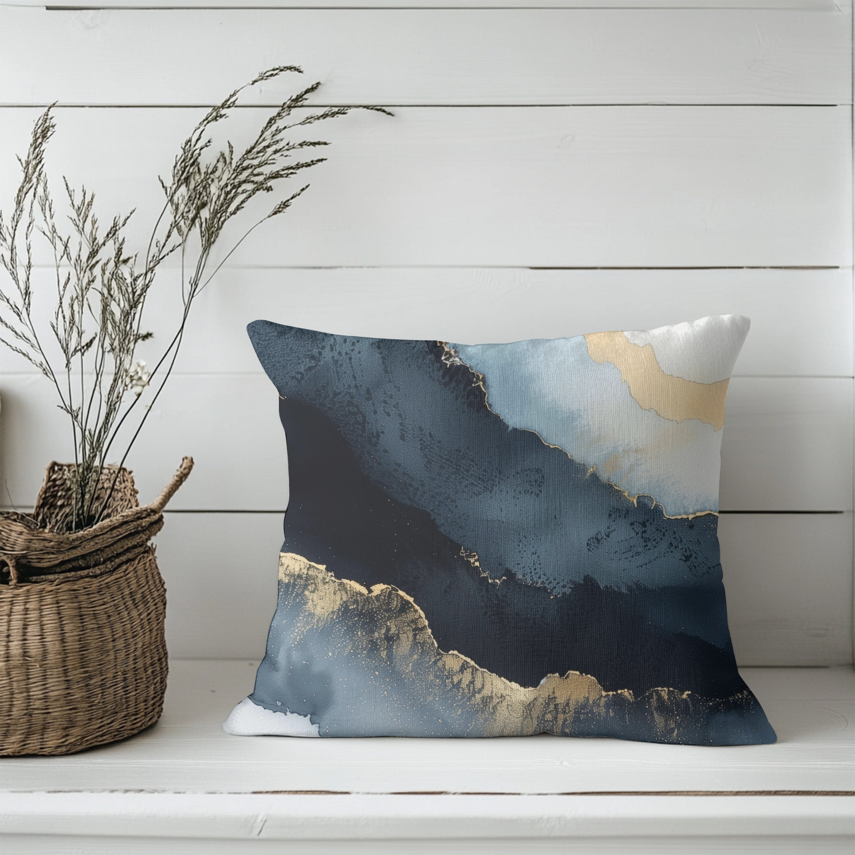 a white wooden shelf with a decorative pillow featuring a blue and gold abstract design, a woven basket with dried plants, and a woven basket with a plant.