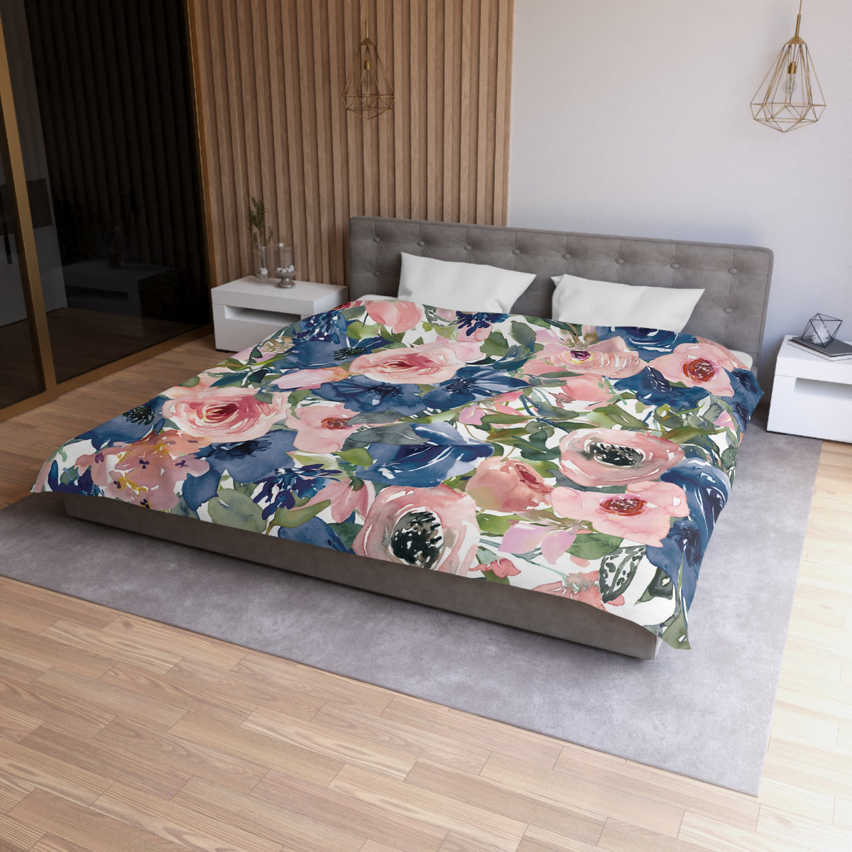 a bedroom with a large bed covered in a floral print comforter. The bed is positioned against a wall with a wooden panel. The room has a wooden floor and a white nightstand on the left side of the bed. There is also a pendant light hanging above the bed.
