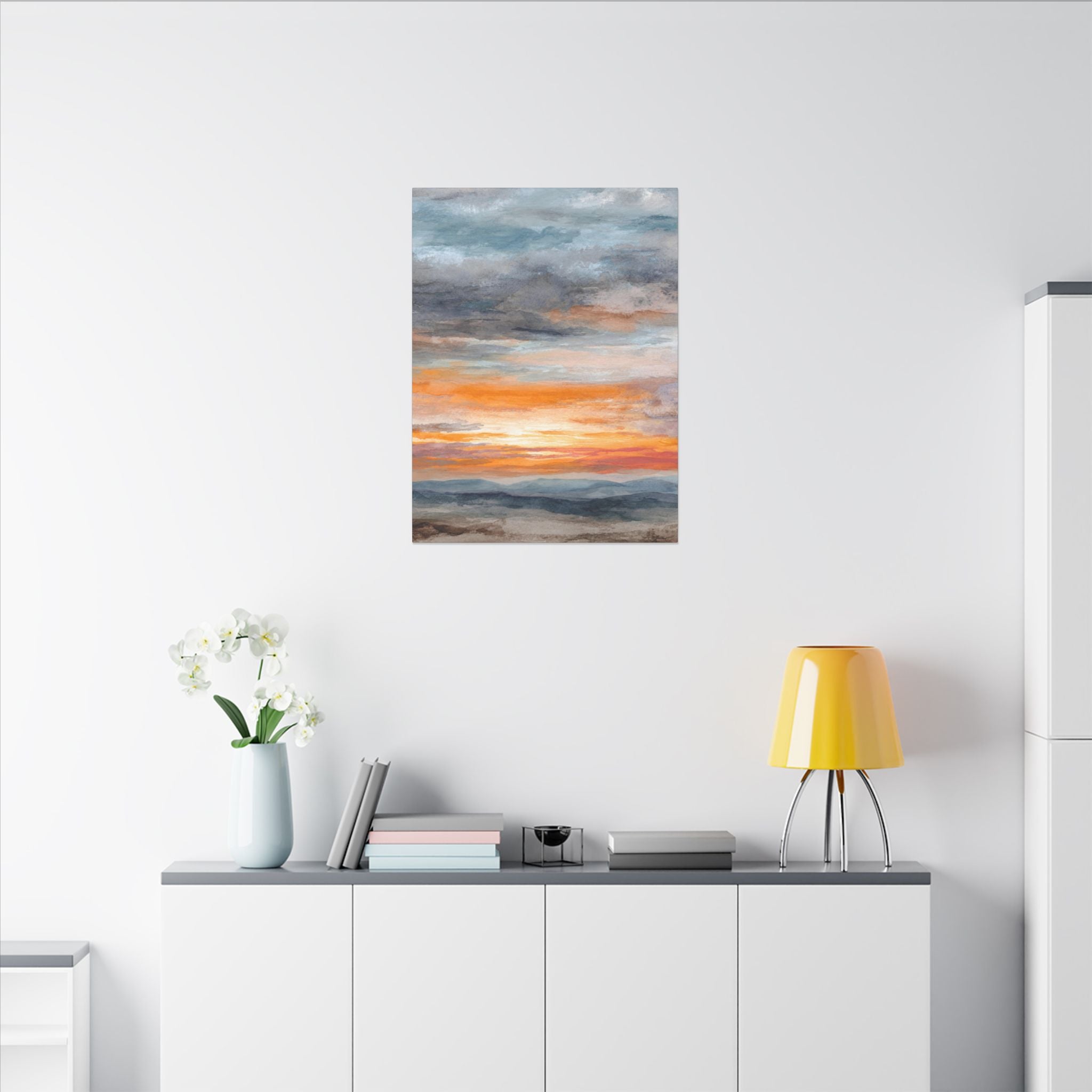 a white wall with a framed painting depicting a sunset over a beach. Below the painting, there is a white shelf with a vase of white flowers, a stack of books, and a yellow lamp.