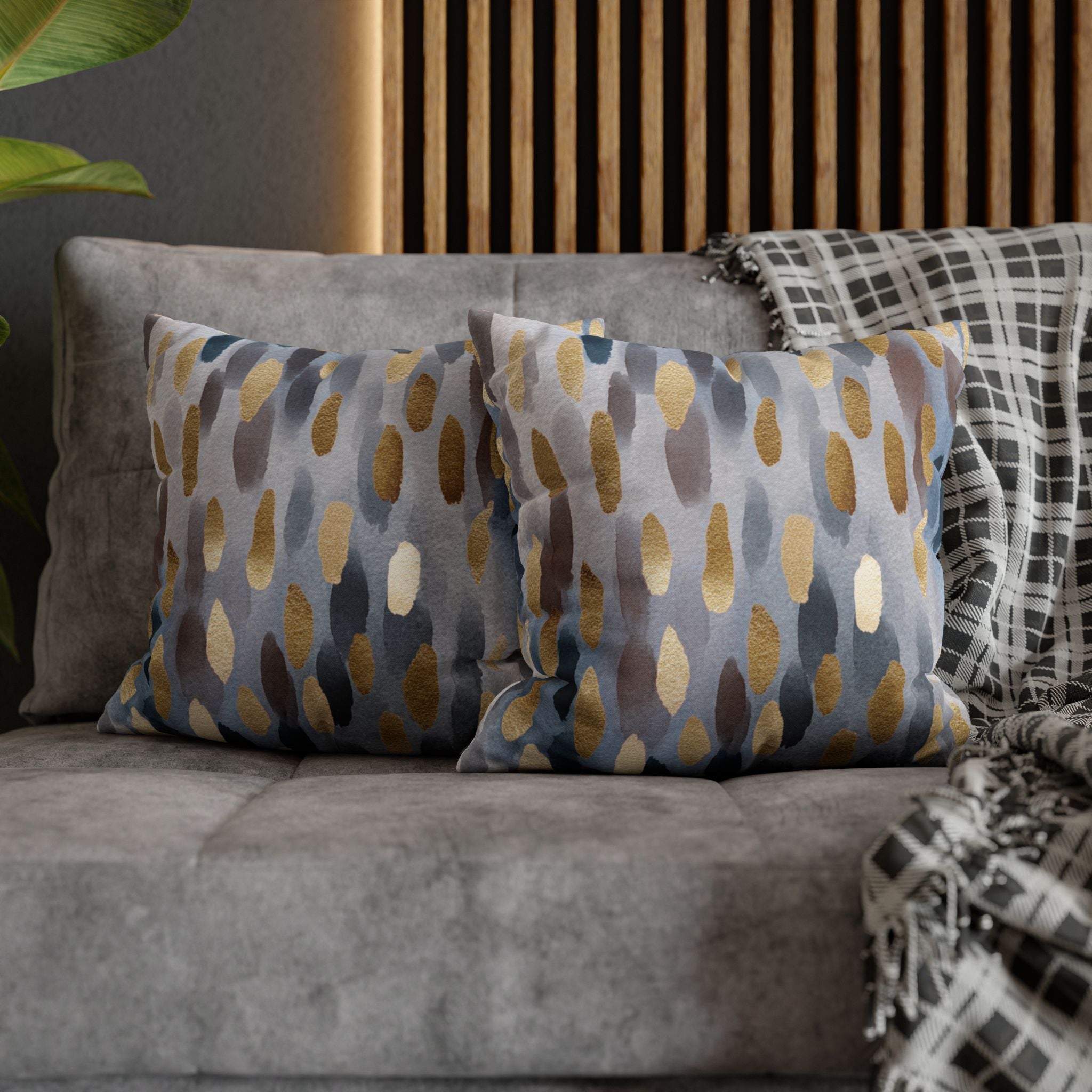 a gray couch with two throw pillows on it, one with a pattern of blue and gold dots and the other with a pattern of gray and white dots. The pillows are placed on a gray couch, and there is a wooden wall and a plant in the background.