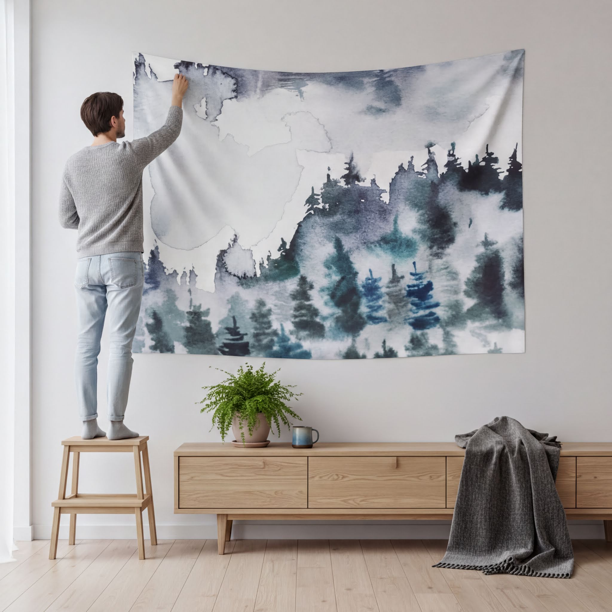 A man is standing on a stool and painting a large tapestry depicting a forest scene on a wall in a room.
