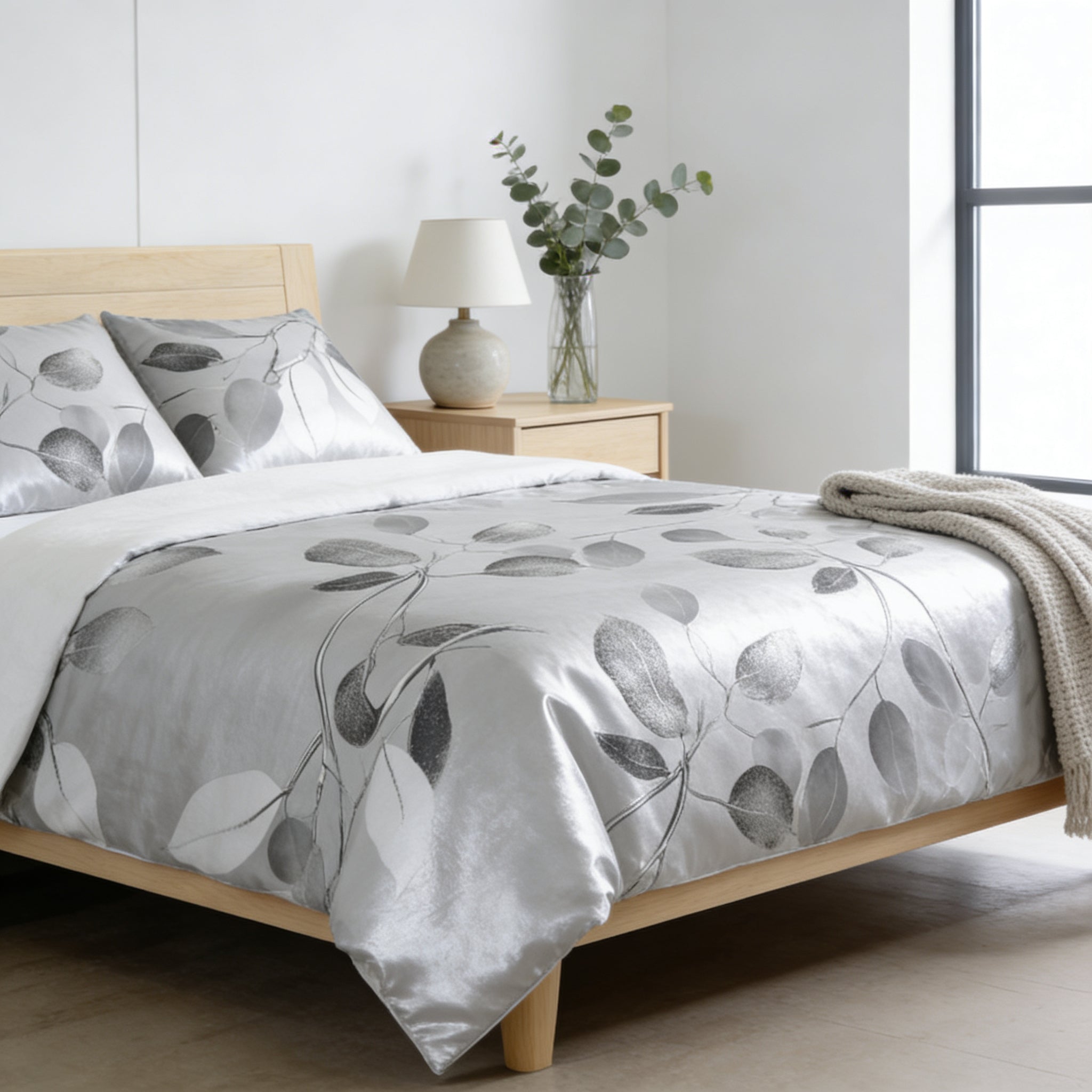 a serene and modern bedroom with a large bed adorned with a gray and white leaf-patterned comforter and pillows. The bed is positioned against a white wall, and a wooden nightstand with a lamp and a vase of greenery is located next to it. The room has a clean and minimalist aesthetic, with a large window providing natural light.