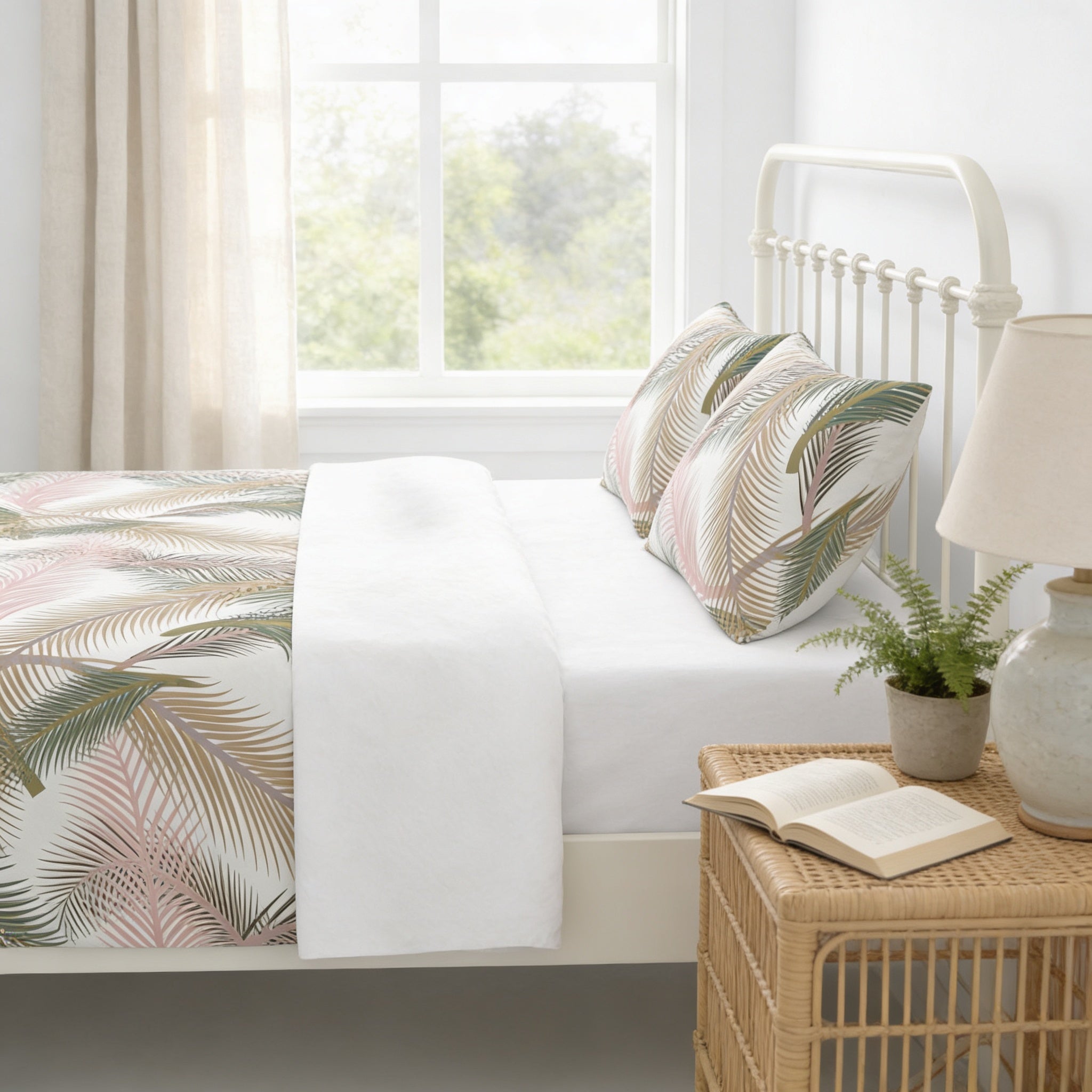 A cozy bedroom with a white metal headboard, a white bedspread with a tropical leaf pattern, and a wicker side table with a lamp and an open book.