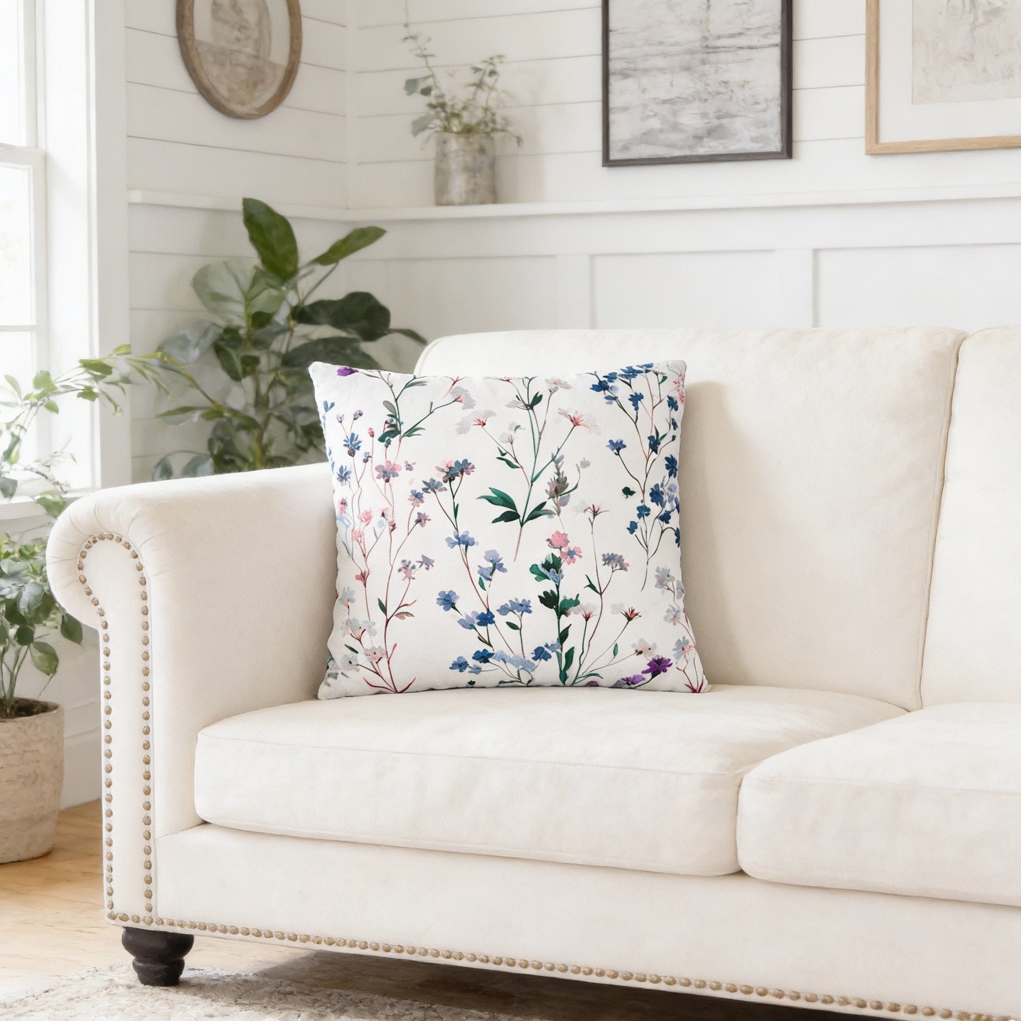 a white sofa with a floral throw pillow on it, placed in a room with a window and some plants.