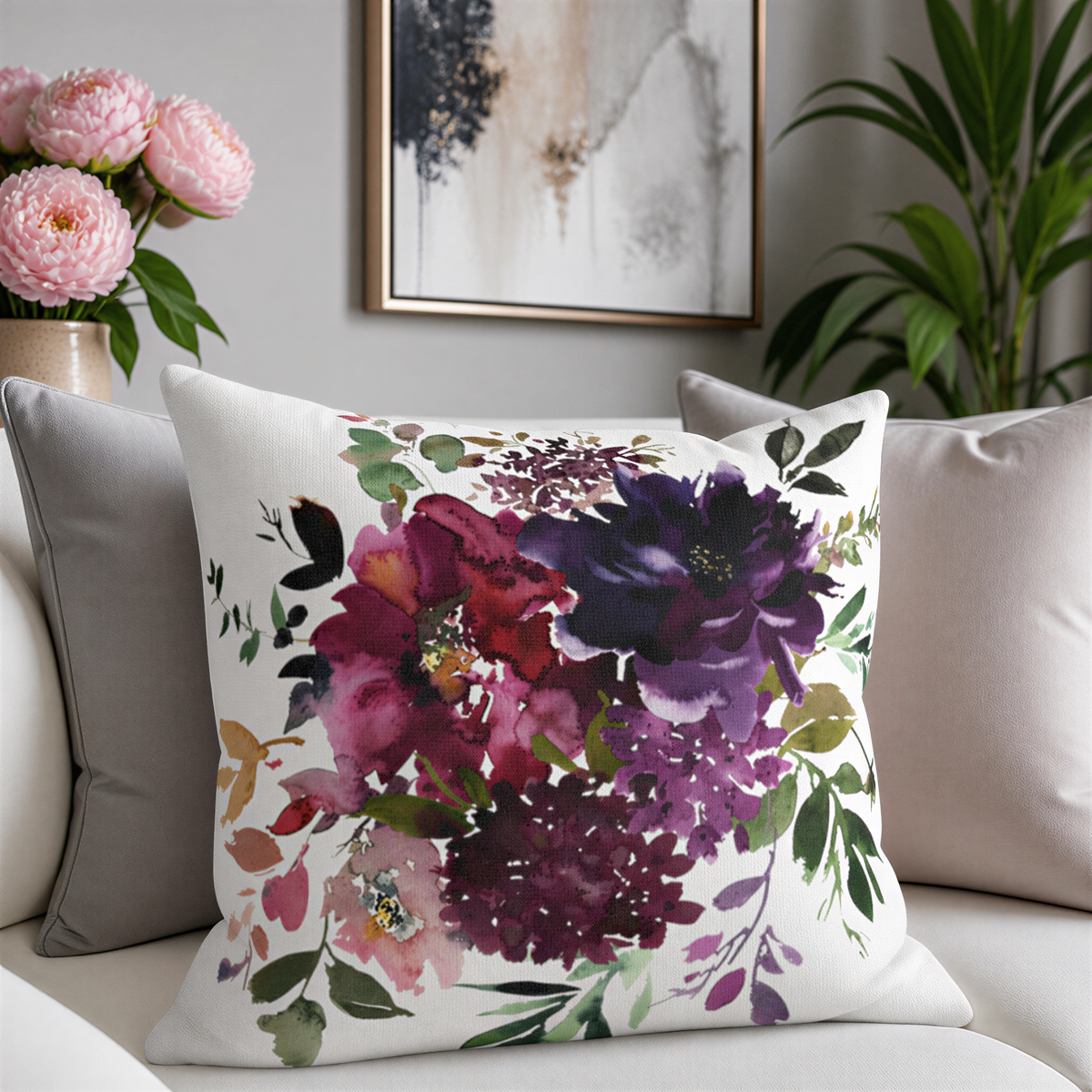 a white couch with a decorative throw pillow featuring a floral design in shades of purple and red. The pillow is placed on the couch, and there is a vase of pink flowers nearby. The background includes a framed picture and a potted plant, creating a cozy and inviting atmosphere.
