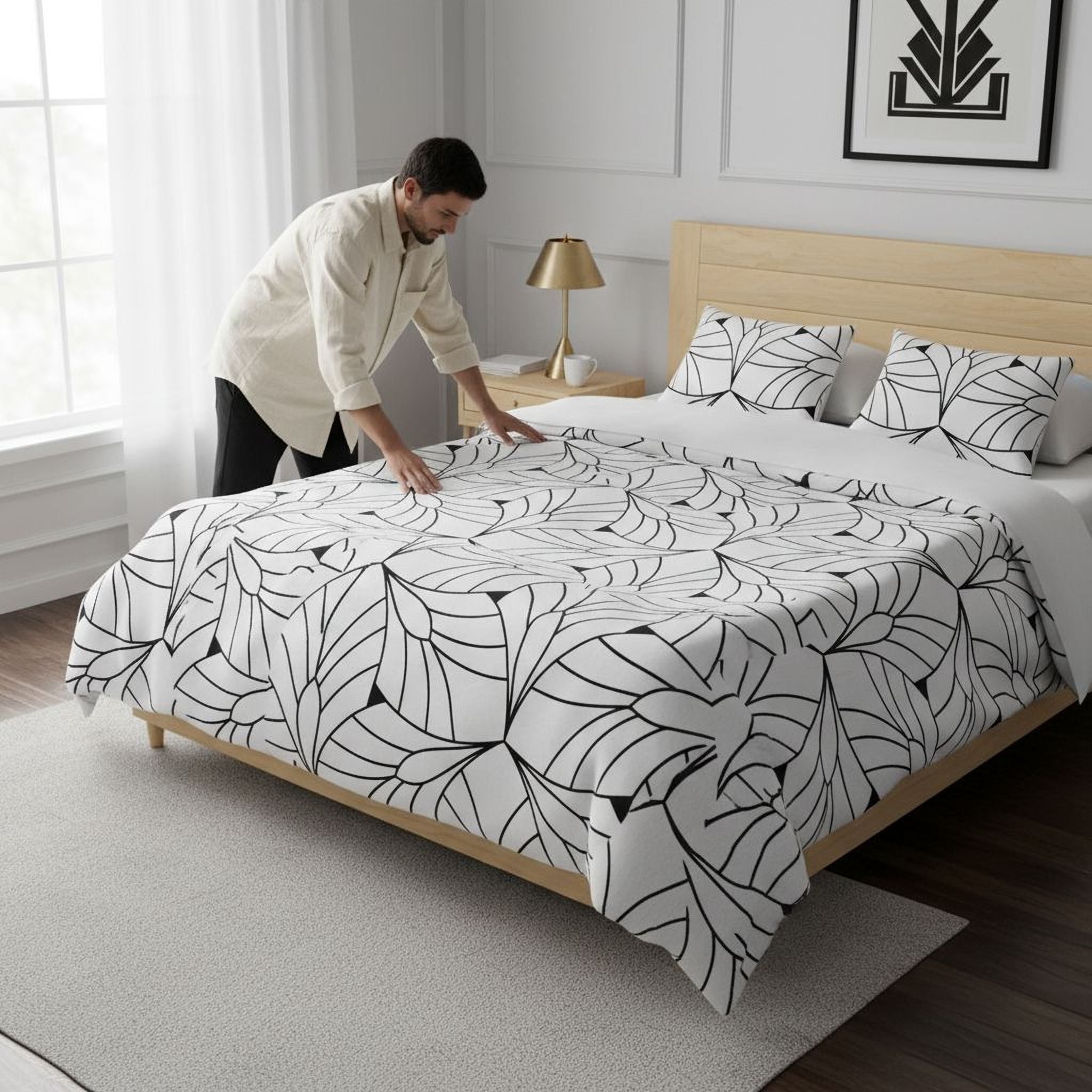 A man is arranging a bed with a black and white leaf-patterned comforter and pillows in a bedroom.