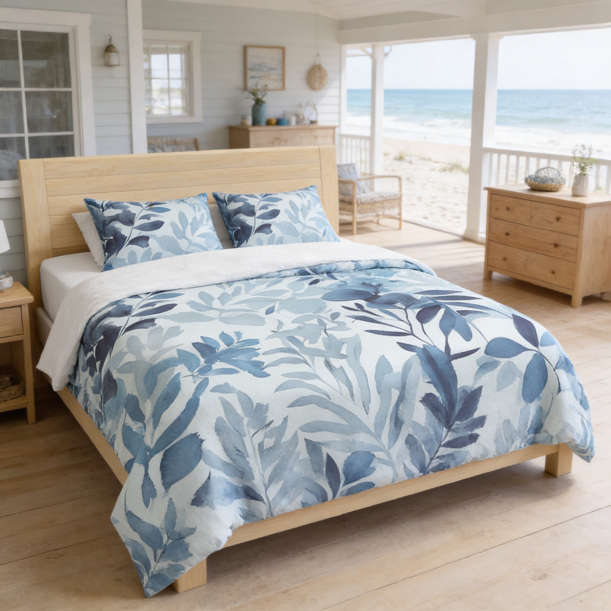 a serene bedroom with a large bed covered in a blue and white leaf-patterned comforter, situated on a wooden floor. The room has a porch with a view of the ocean, and there are two chairs and a dresser nearby. The overall atmosphere of the room is calm and inviting, with the natural elements of the ocean and the wooden furniture complementing each other.