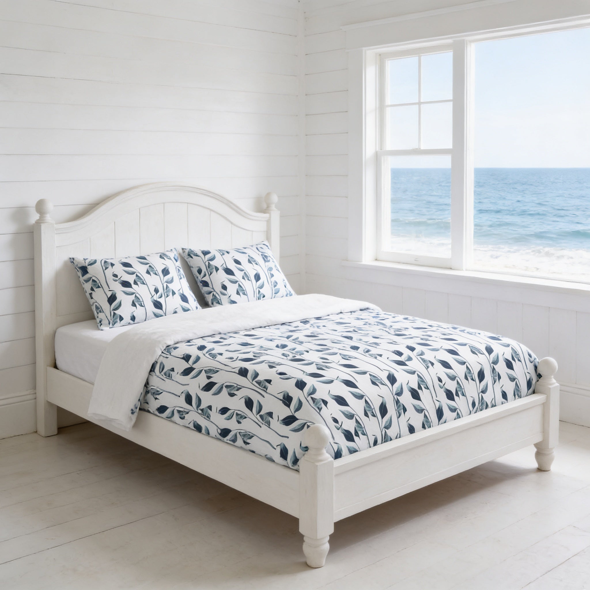 A white wooden bed with a blue and white leaf patterned comforter and pillows, situated in a bright and airy room with a large window overlooking the ocean.