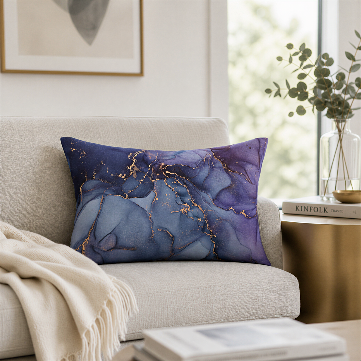 a living room with a white sofa, a decorative pillow with a marble-like pattern in shades of blue and purple, and a framed picture on the wall. There is also a vase with greenery on a side table, and a stack of books on a coffee table.