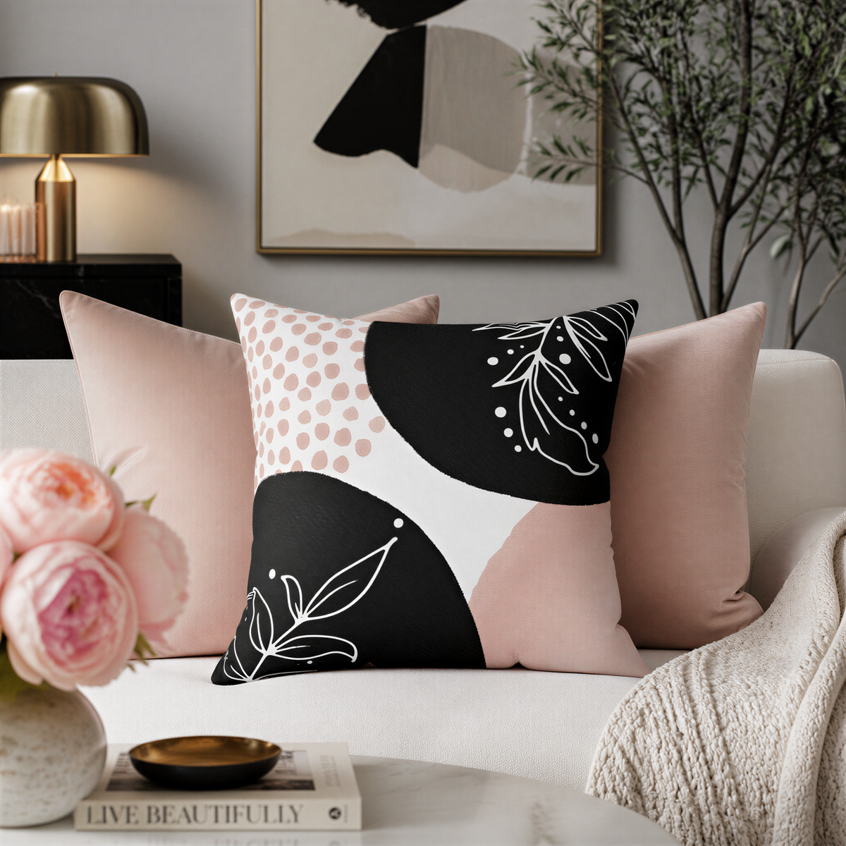 a living room with a white sofa adorned with three throw pillows, one of which has a black and white polka dot pattern and another with a floral design. A framed abstract painting hangs on the wall, and a vase of pink roses is placed on the coffee table. A book titled "Live Beautifully" is also visible on the table.