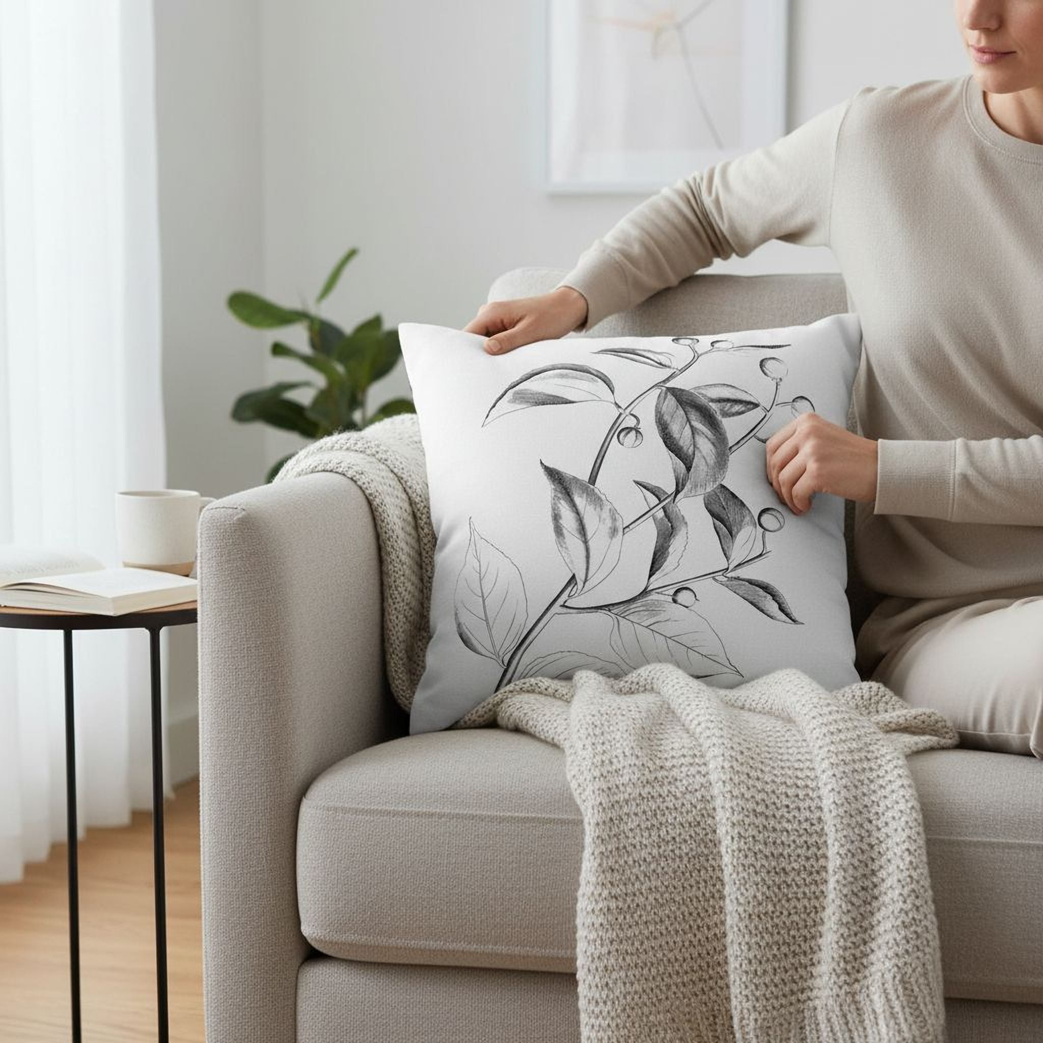 A person is sitting on a couch, adjusting a white pillow with a black and white leaf design.