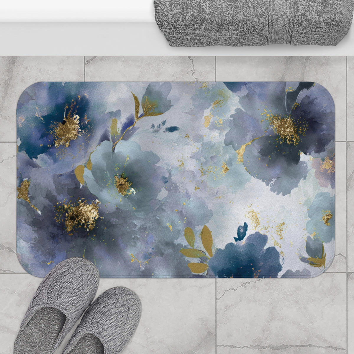 a bathroom mat with a floral design, including blue and gold flowers, placed on a tiled floor. There are also a pair of gray slippers placed next to the mat.