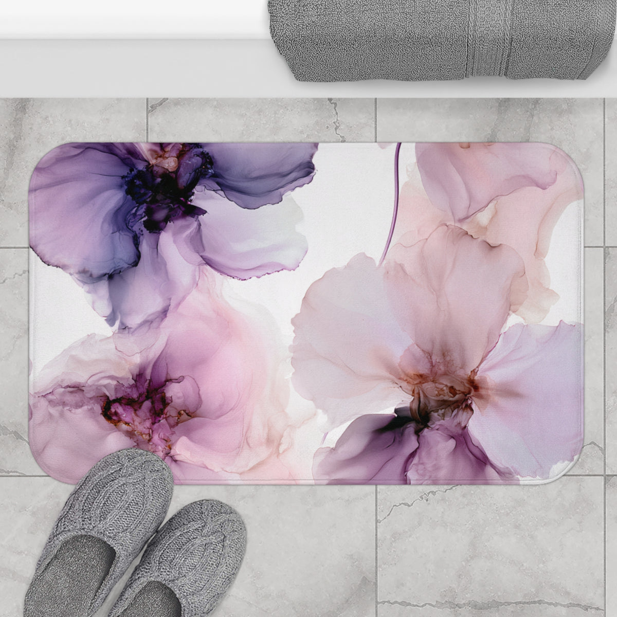 a bathroom mat with a floral design, including purple and pink flowers, placed on a tiled floor. There are also gray slippers and a gray towel visible in the scene.
