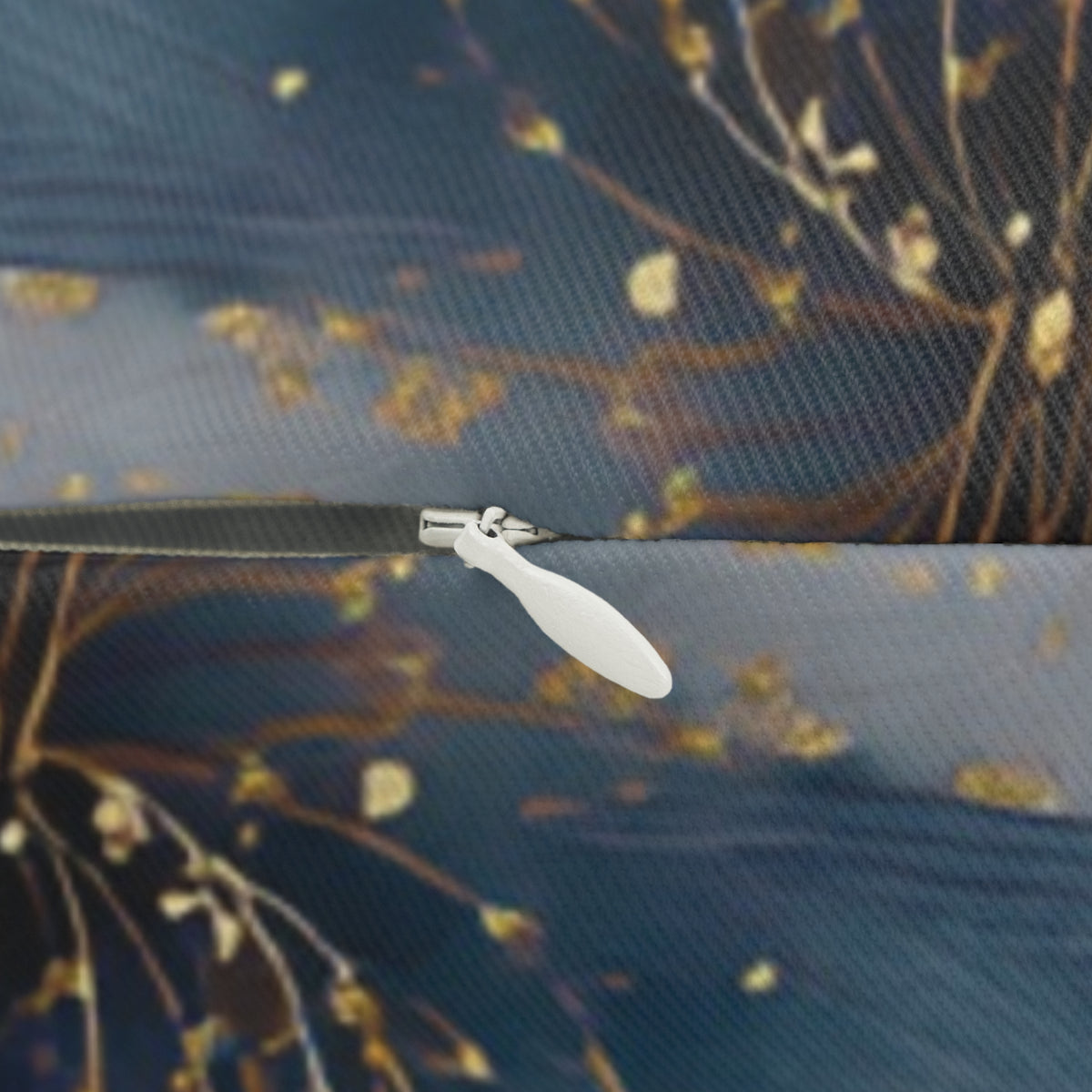 A close-up view of a zipper on a fabric, with a blurred background featuring a pattern of leaves and branches.