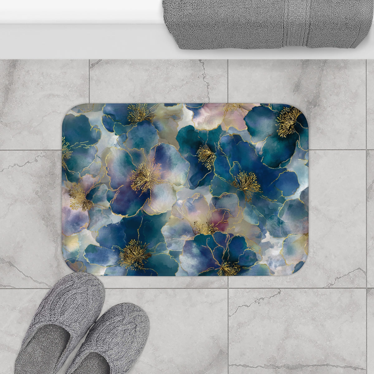 a bathroom floor with a decorative floral pattern on the floor mat, gray slippers placed next to it, and a rolled-up towel on the countertop.
