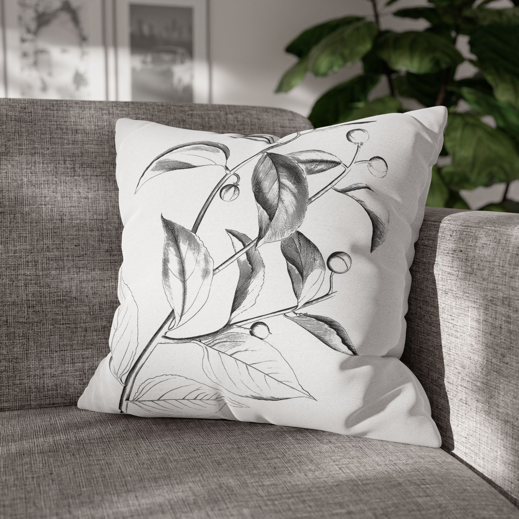 A white throw pillow with a black and white leaf design sits on a gray couch, with a plant visible in the background.