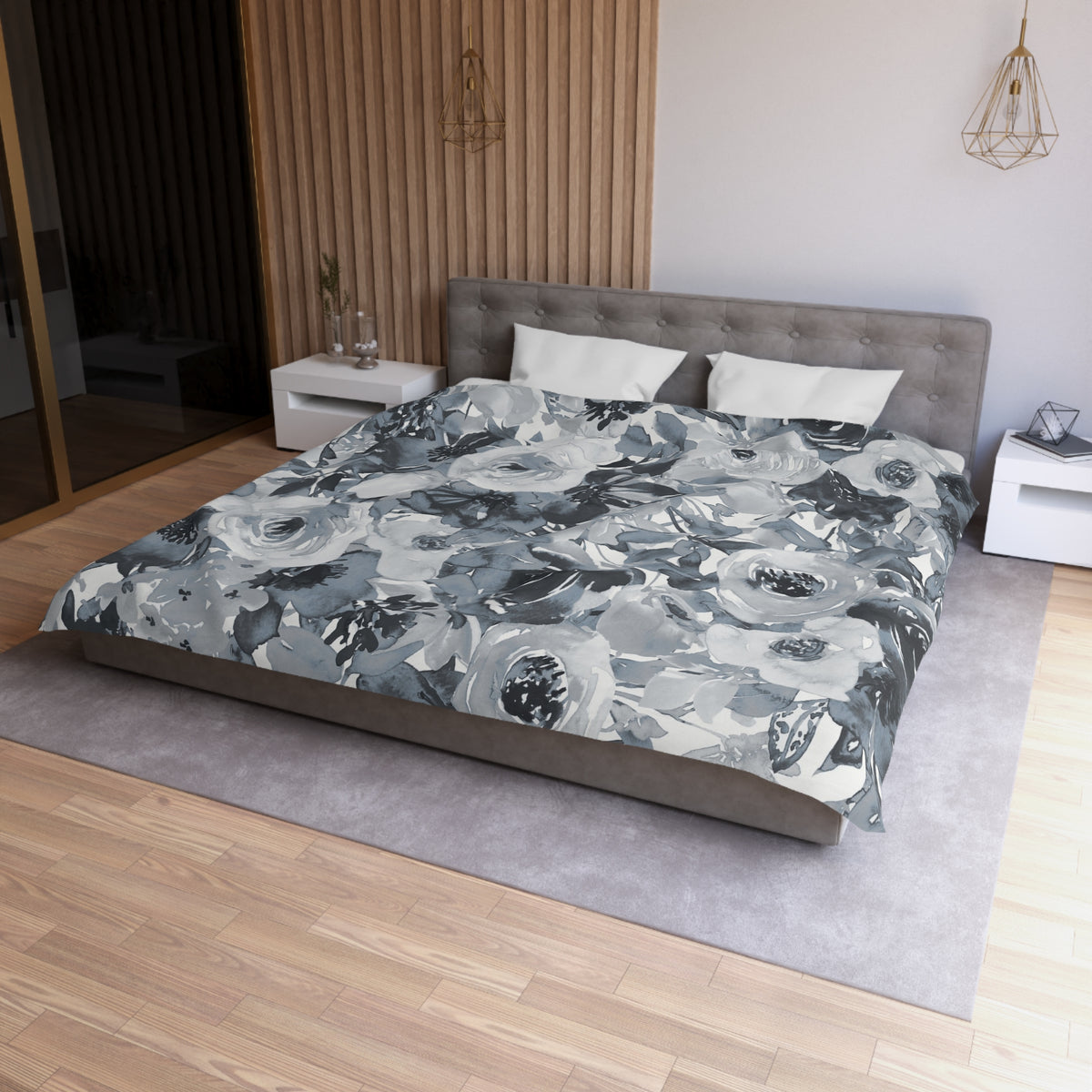 a modern bedroom with a large bed covered in a gray and white floral comforter. The bed is positioned against a wall with a wooden panel. The room also features a nightstand on the left side and a pendant light hanging above the bed.