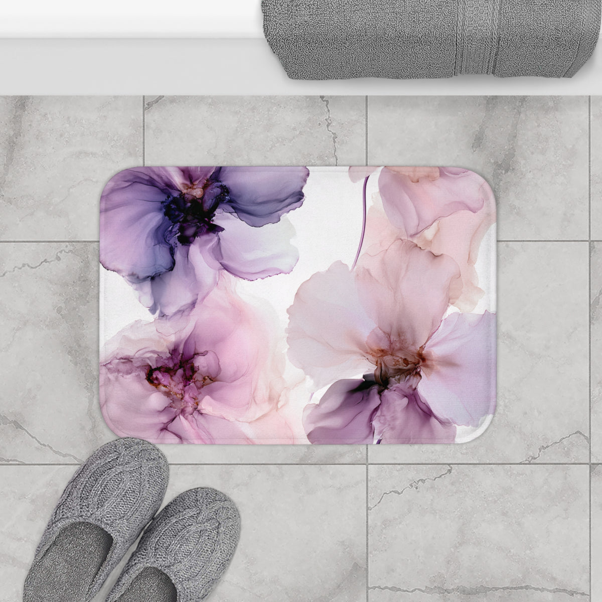 a bathroom floor with a floral patterned bath mat, gray slippers, and a rolled-up gray towel.