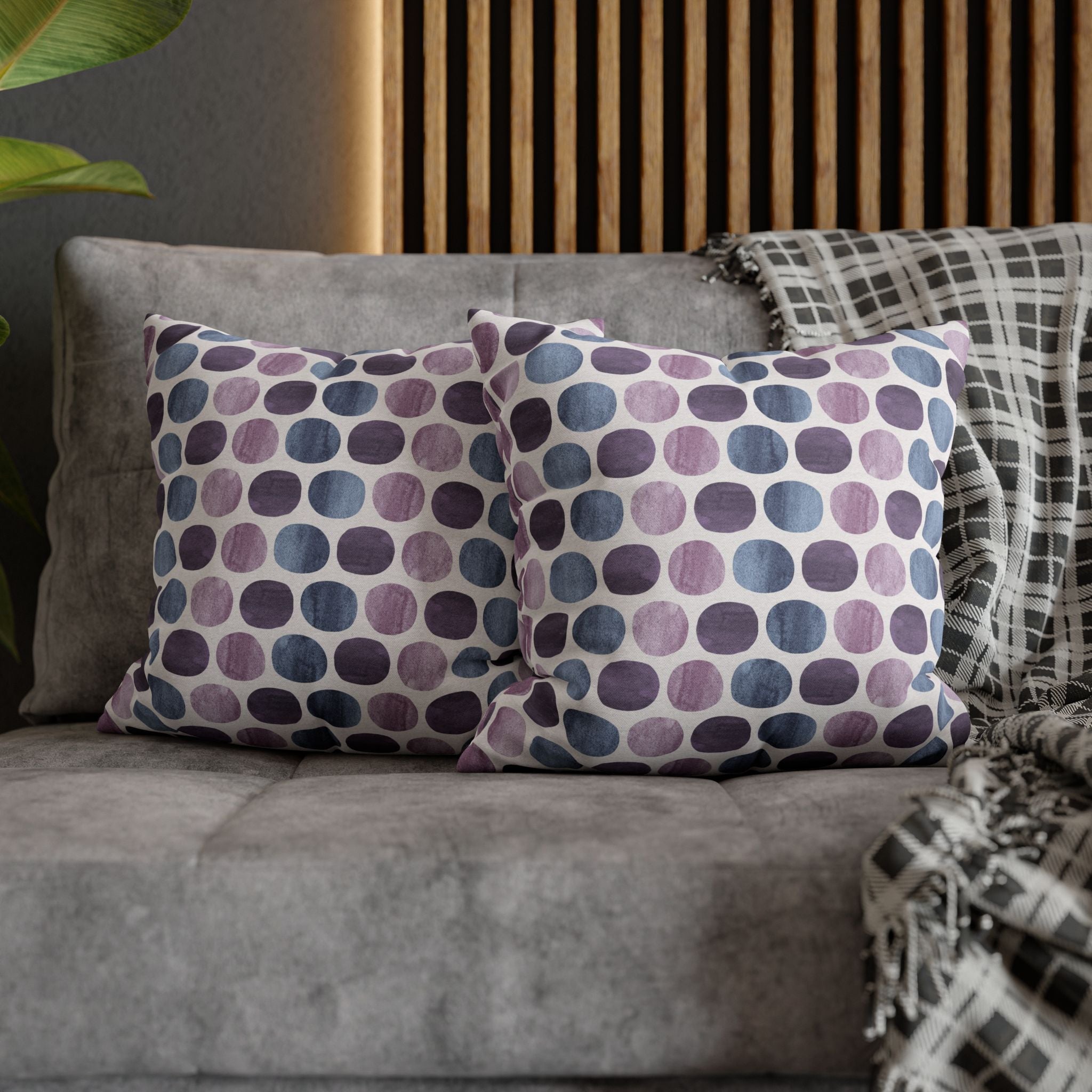 a gray couch with two throw pillows on it. The pillows have a polka dot pattern in shades of purple and blue. The couch is positioned against a wooden wall, and there is a plant visible in the background.