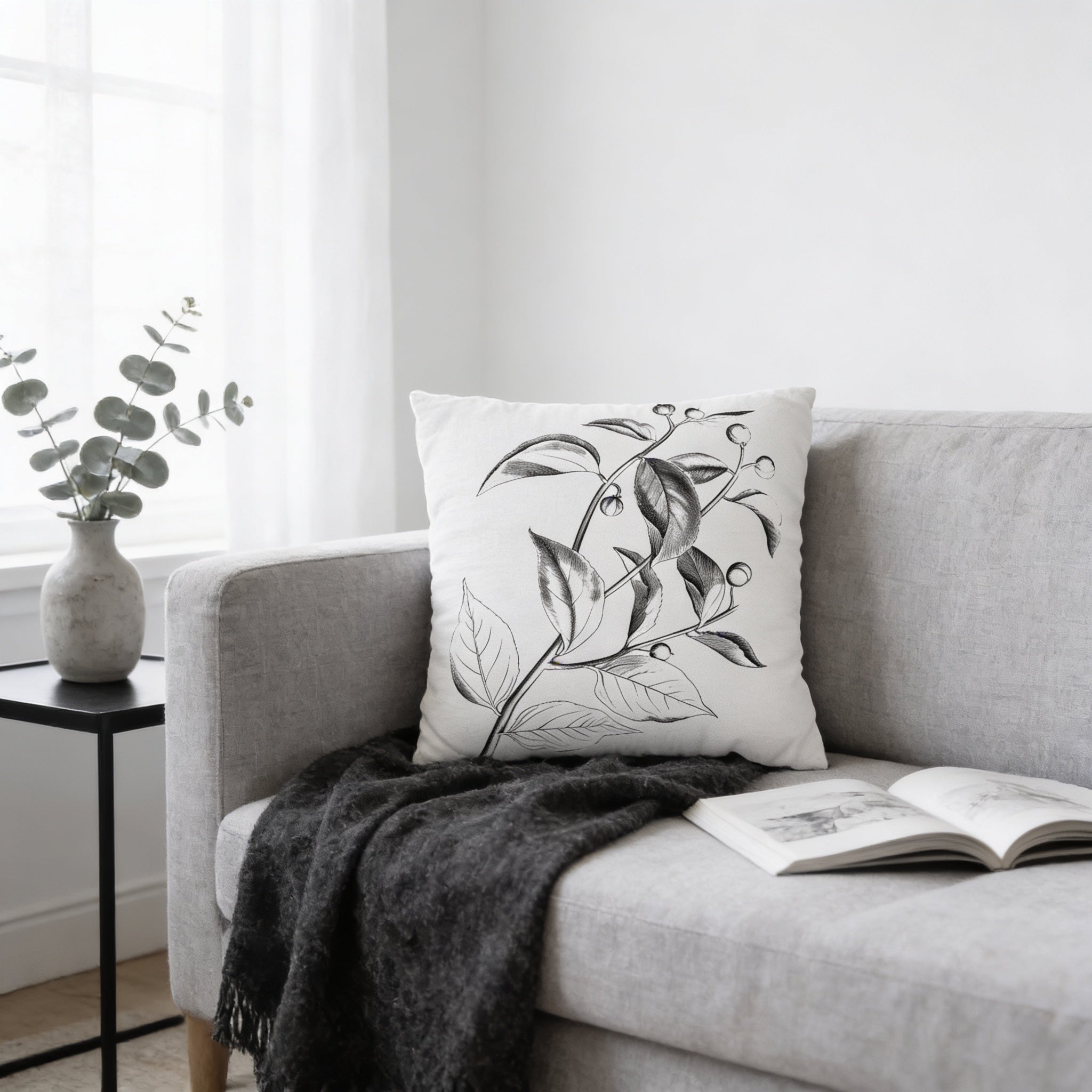 a cozy living room with a gray sofa, a white pillow with a black leaf design, a black blanket, and an open book on the sofa.