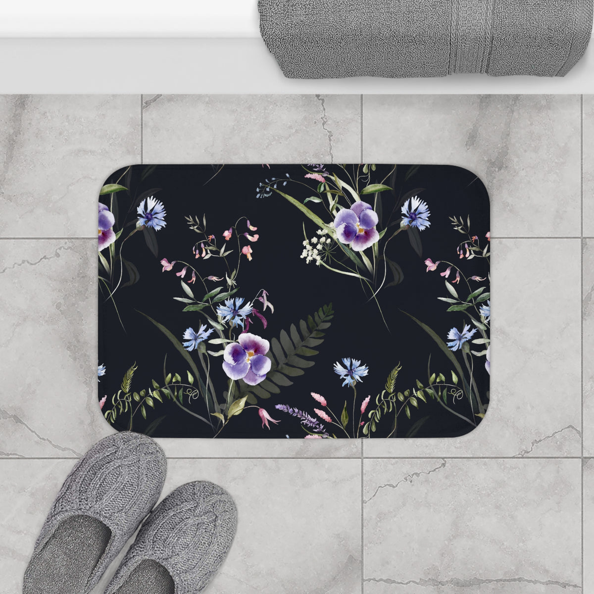 a bathroom floor with a floral patterned bath mat, gray slippers, and a rolled-up towel.