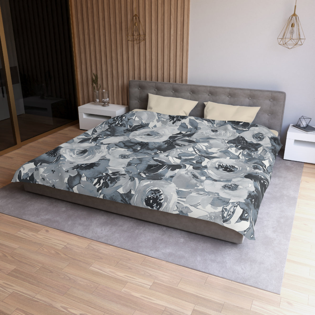 a modern bedroom with a large bed covered in a gray and white floral comforter. The bed is positioned against a wall with a wooden panel. The room also features a nightstand on the left side and a pendant light hanging above the bed.