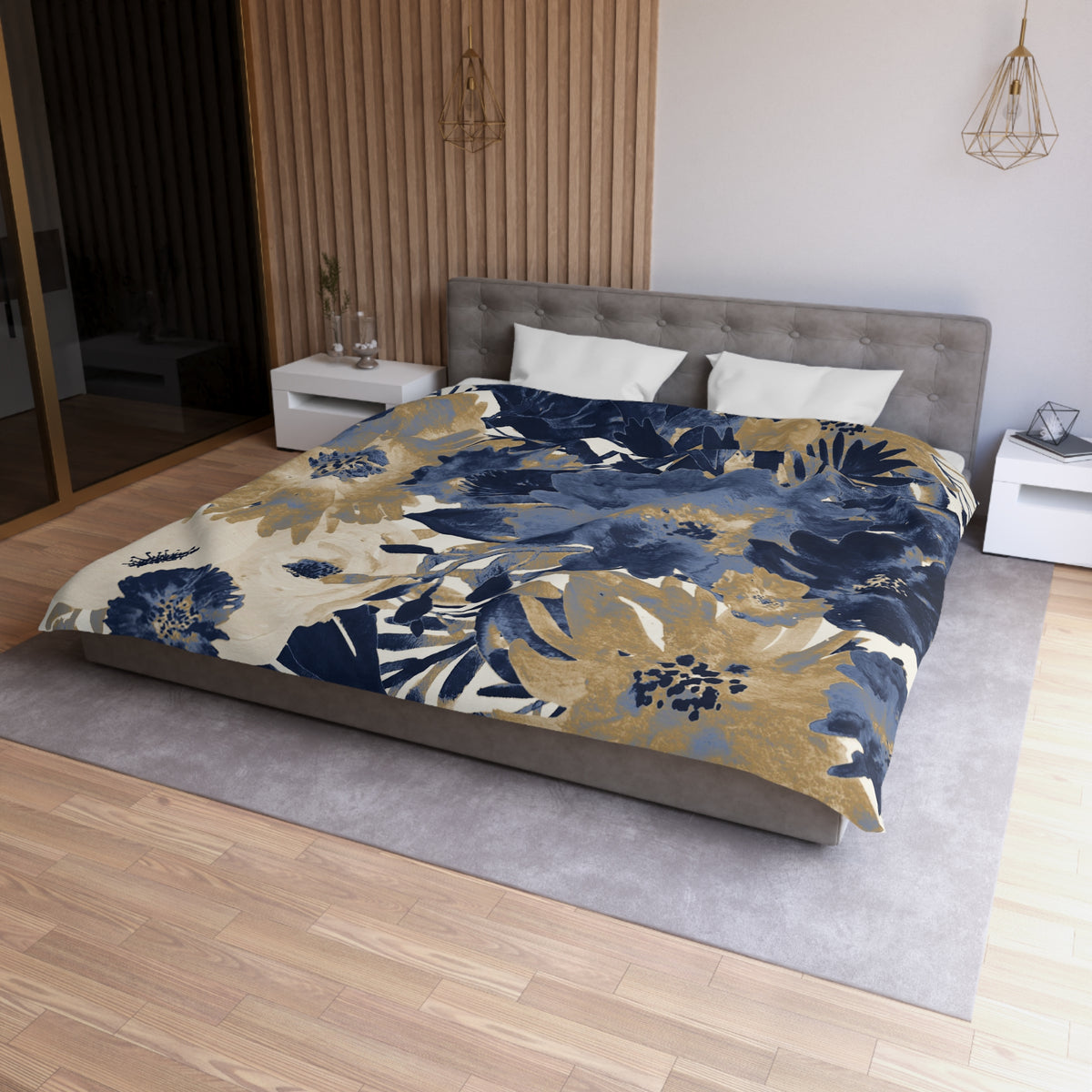 a modern bedroom with a large bed covered in a blue and gold floral comforter. The bed is positioned against a wall with a wooden panel. The room also features a nightstand, a pendant light, and a vase on a surface.