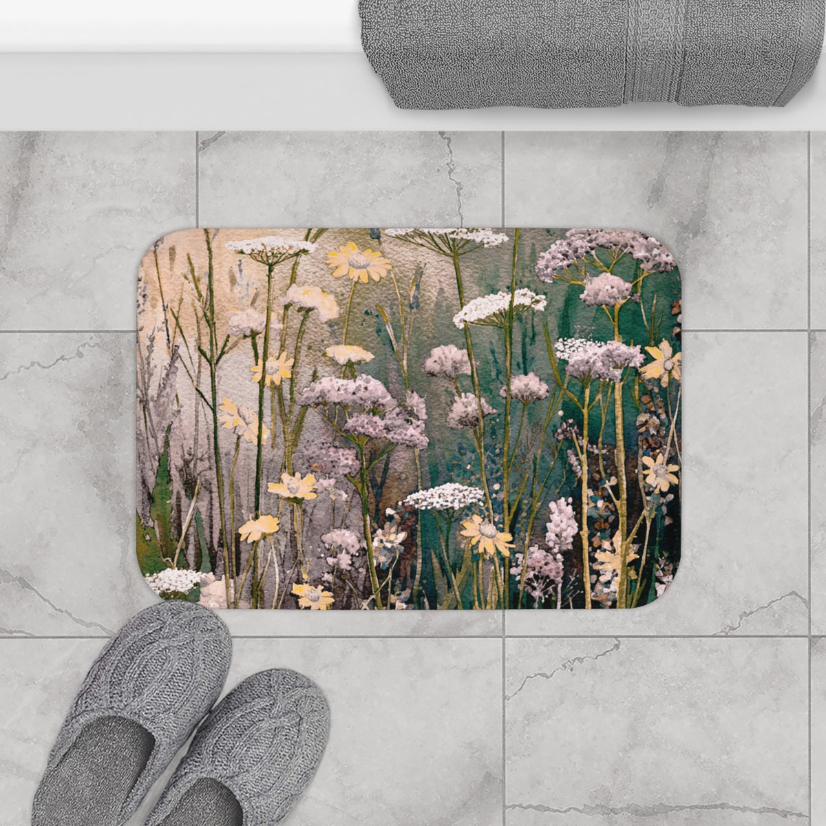 a bathroom floor with a rectangular bath mat featuring a floral design, a pair of gray slippers, and a rolled-up gray towel.