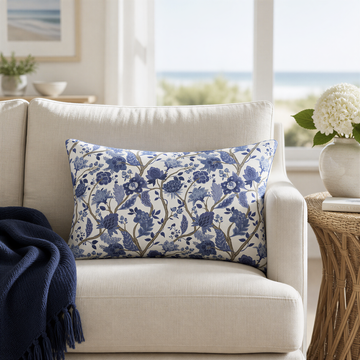 a cozy living room with a beige sofa adorned with a blue and white floral throw pillow. A white vase with white flowers is placed on a side table, and a blue blanket is draped over the arm of the sofa. The room has a window overlooking the ocean, creating a serene and relaxing atmosphere.