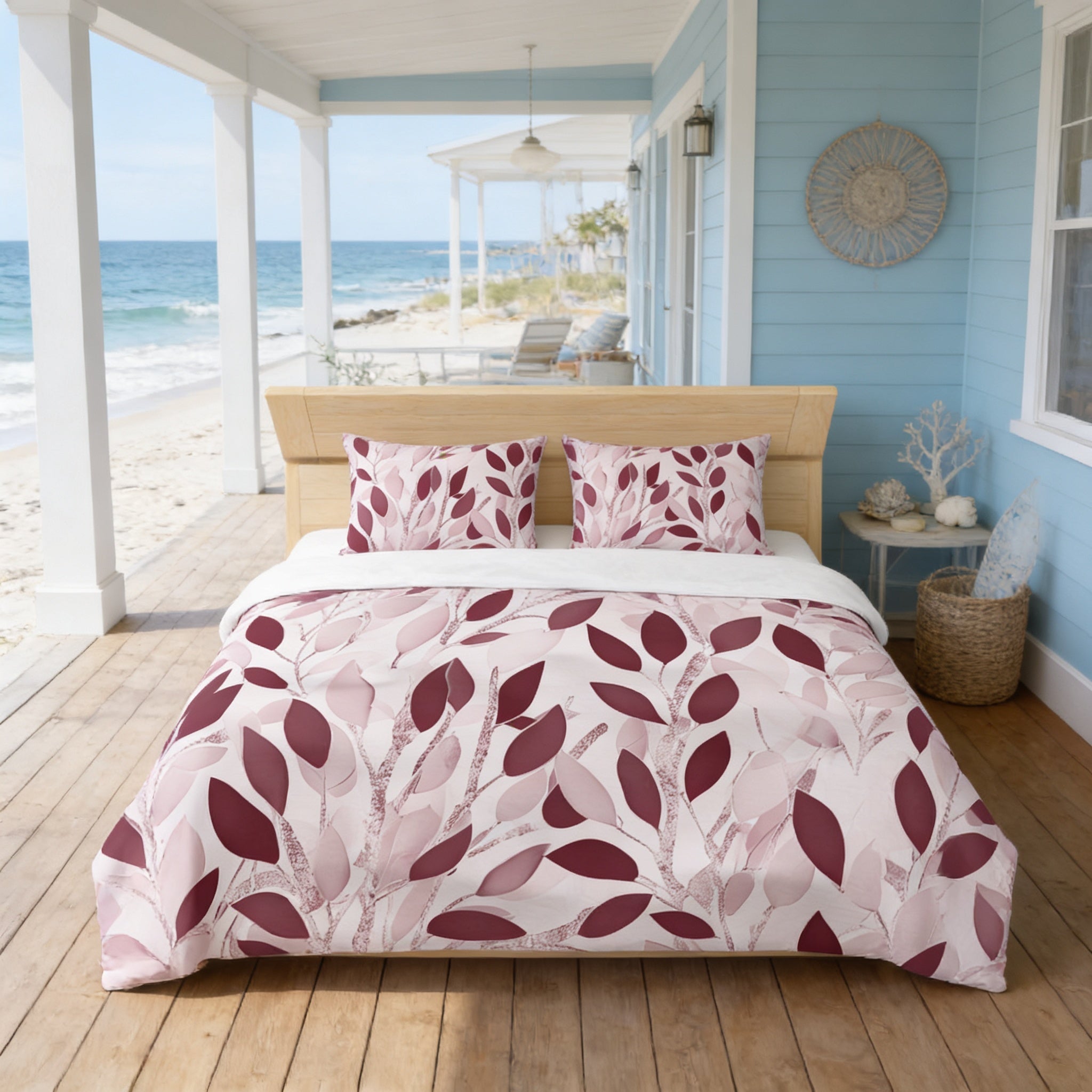 a bedroom with a large bed, a wooden headboard, and a view of the ocean through a covered porch. The bed has a comforter with a leaf pattern in shades of pink and white. The room has a coastal theme, with a wicker basket and a small table nearby. The overall color scheme is a mix of blue, white, and pink.