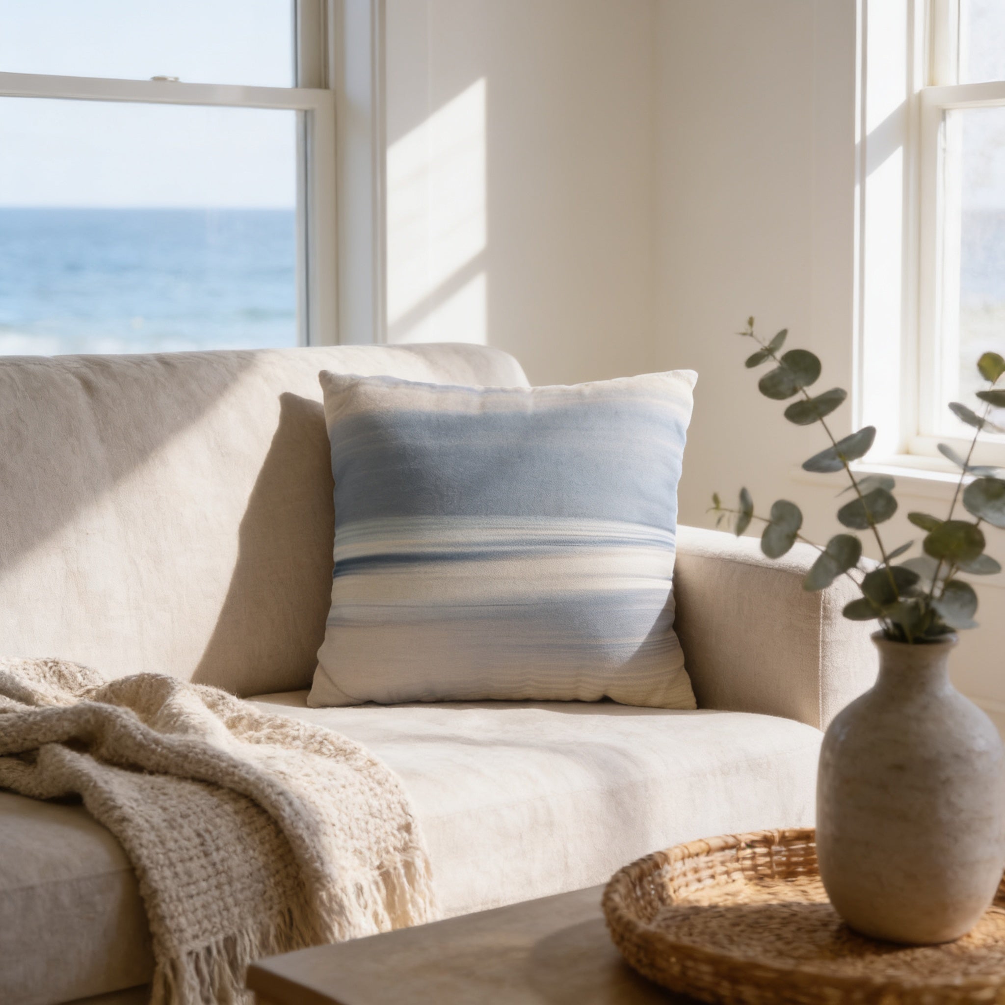 a cozy living room with a beige sofa, a blue and white striped pillow, a woven basket with greenery, and a large window overlooking the ocean.