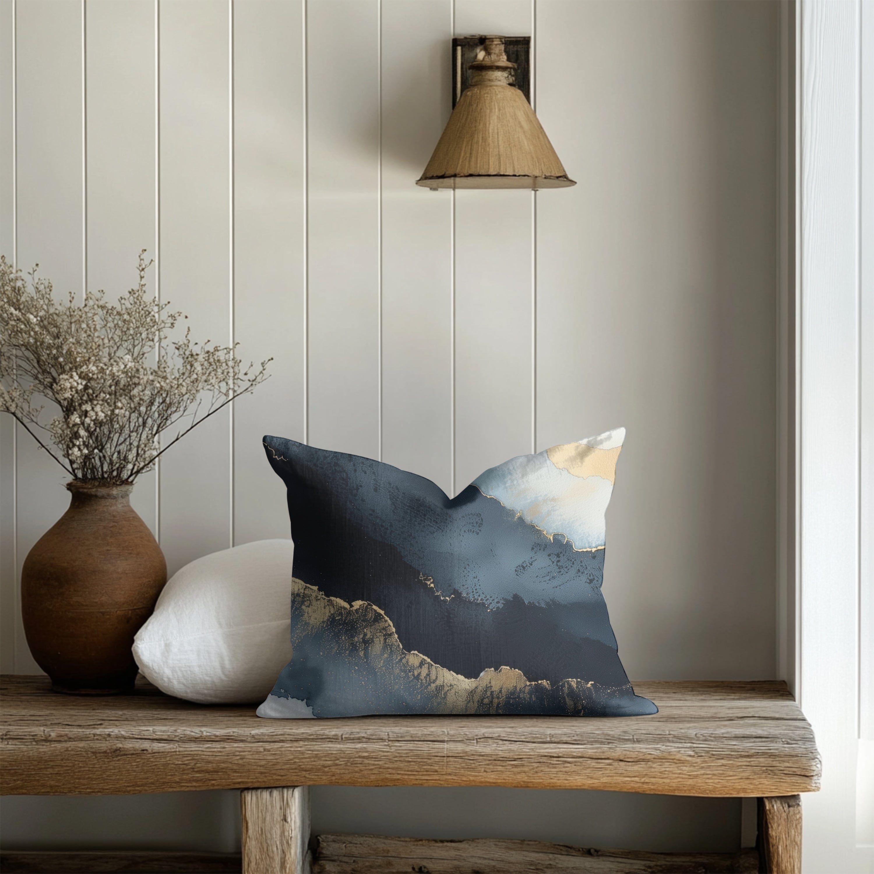 a wooden bench with a decorative pillow on it, placed against a white wall with a light fixture above. There is also a vase of flowers and a potted plant on the bench.