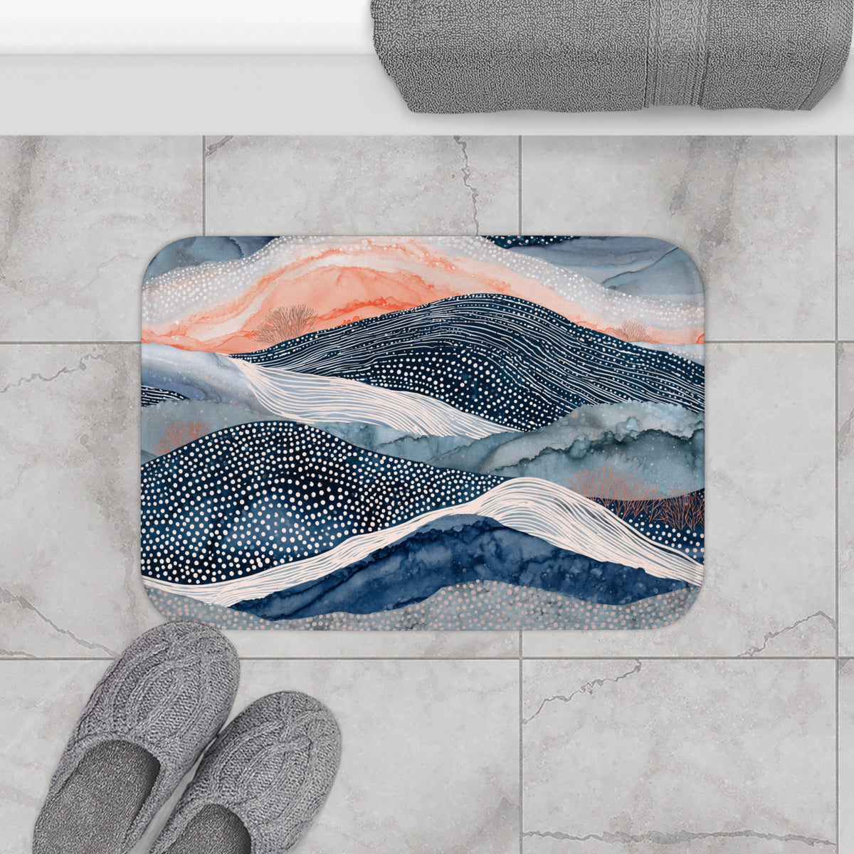 a bathroom floor with a decorative bath mat featuring a mountain landscape design in shades of blue, orange, and white. Next to the bath mat are a pair of gray slippers placed on the floor.