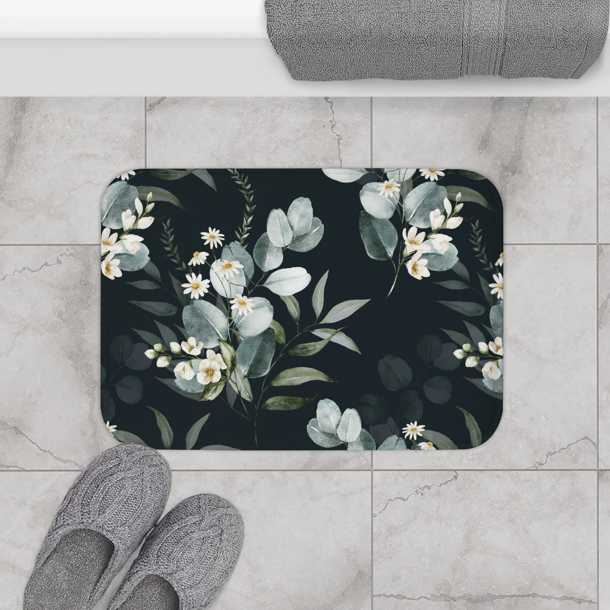 a bathroom floor with a floral patterned bath mat, gray slippers, and a rolled-up towel.
