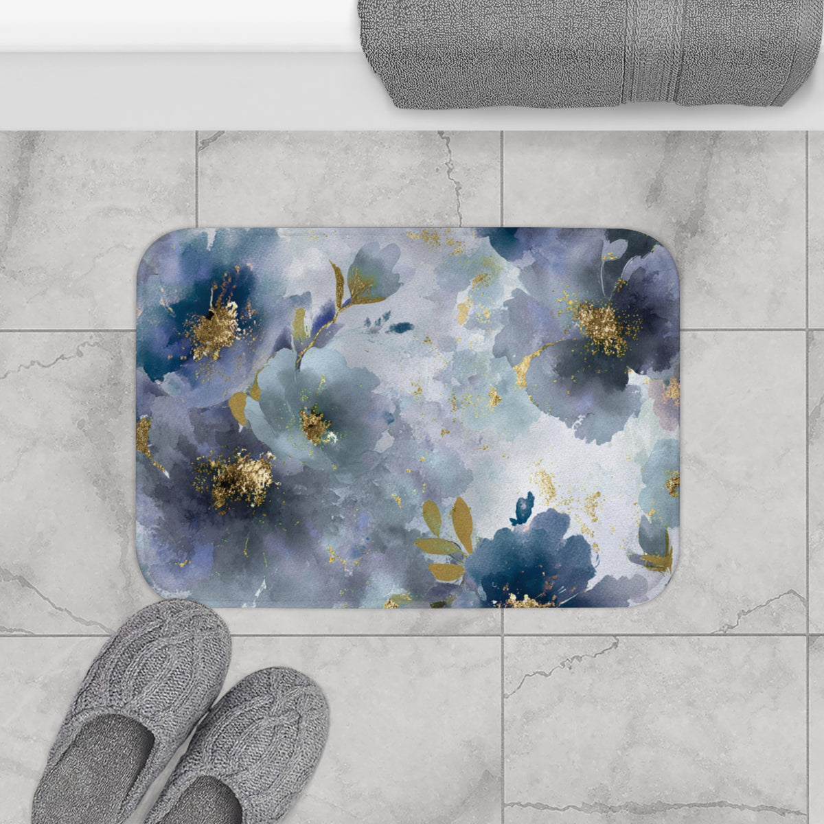 a bathroom floor with a decorative floral pattern on a rectangular bath mat, a pair of gray slippers, and a rolled-up gray towel.