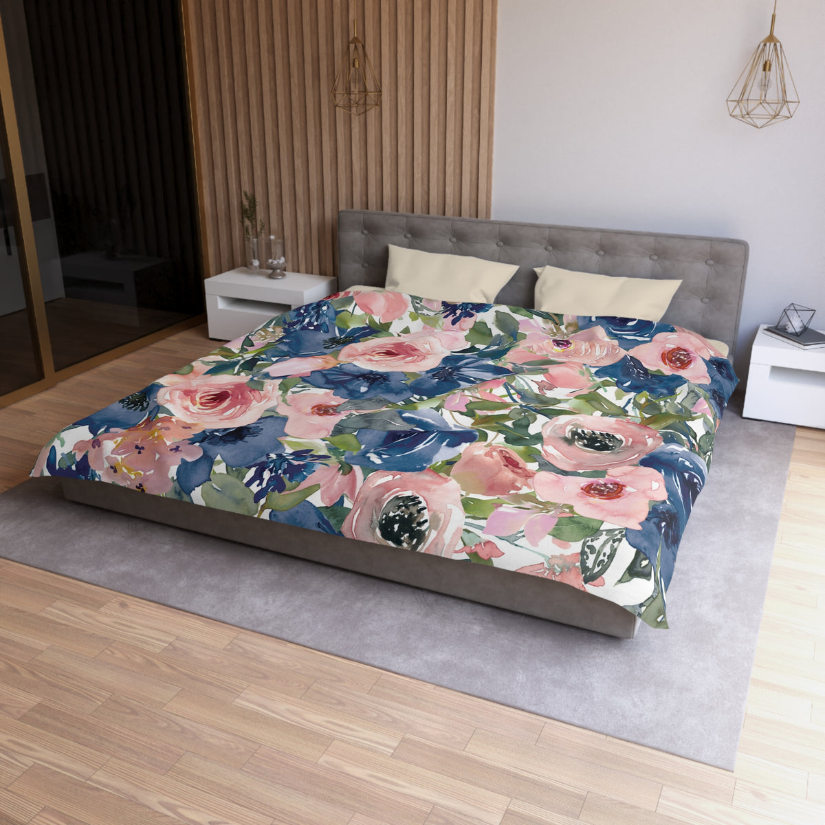 a bedroom with a large bed covered in a floral print comforter. The bed is positioned against a wall with a wooden panel. The room has a wooden floor and a white nightstand on the left side of the bed. There is also a pendant light hanging above the bed.
